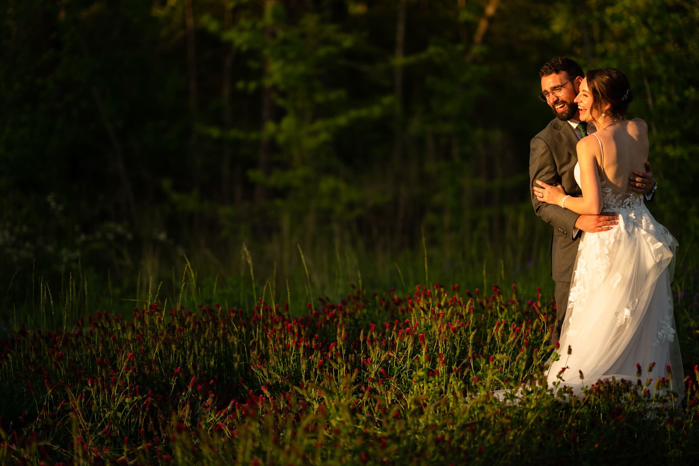 union grove farm wedding photography - kaylyn & jeremy A couple stands in a field of red flowers at Union Grove Farm, surrounded by lush greenery. The bride, in a white dress, smiles and holds the groom in his suit. Warm sunlight illuminates their joyful moment, captured beautifully as a part of their wedding photography against shaded trees.