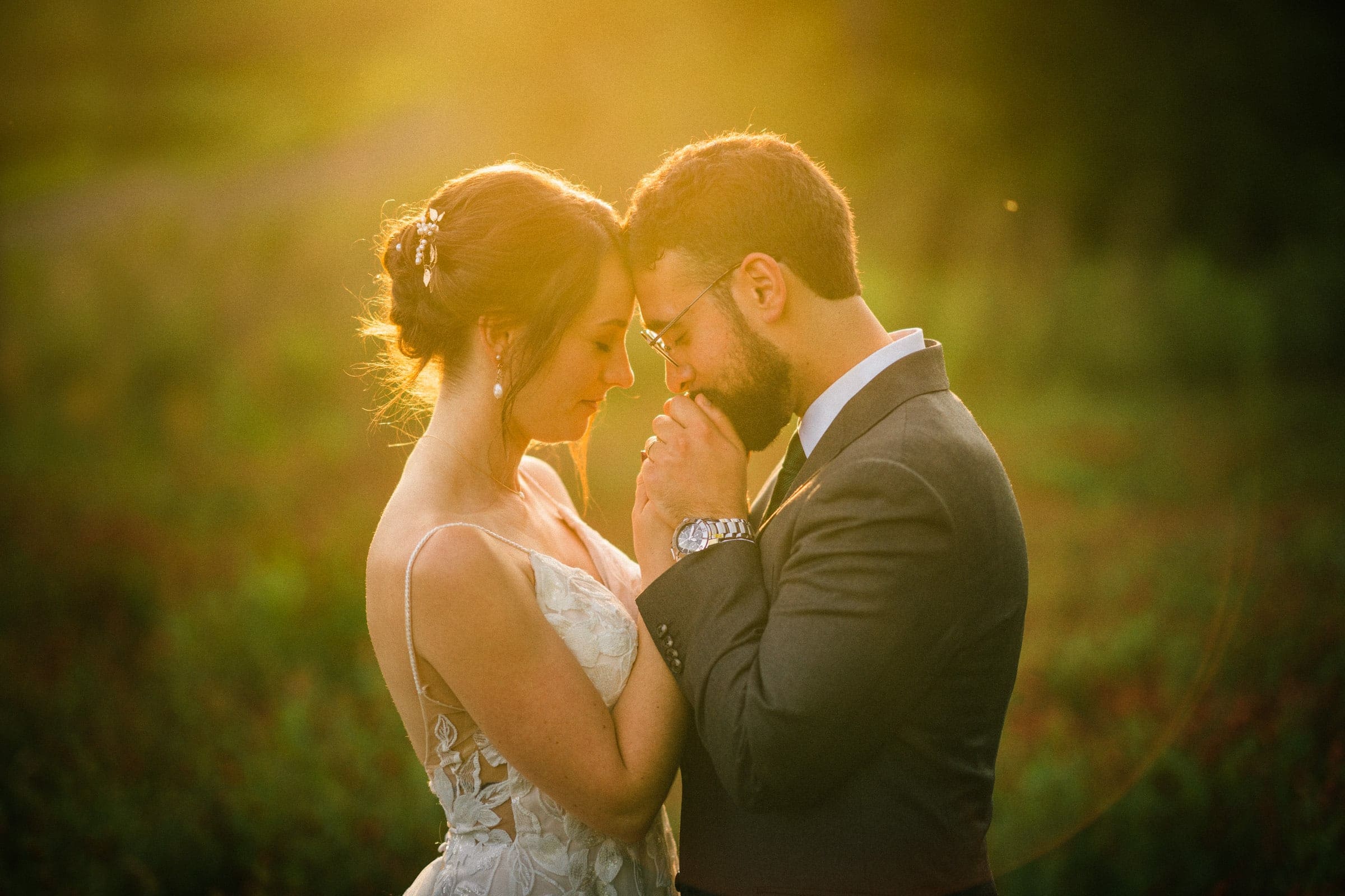 union grove farm wedding photography - kaylyn & jeremy A couple stands closely, gently touching foreheads, surrounded by the warm, golden sunlight of Union Grove Farm. The woman is in a white dress, and the man is in a gray suit. They hold hands, creating an intimate and serene moment captured perfectly through wedding photography in this natural setting.