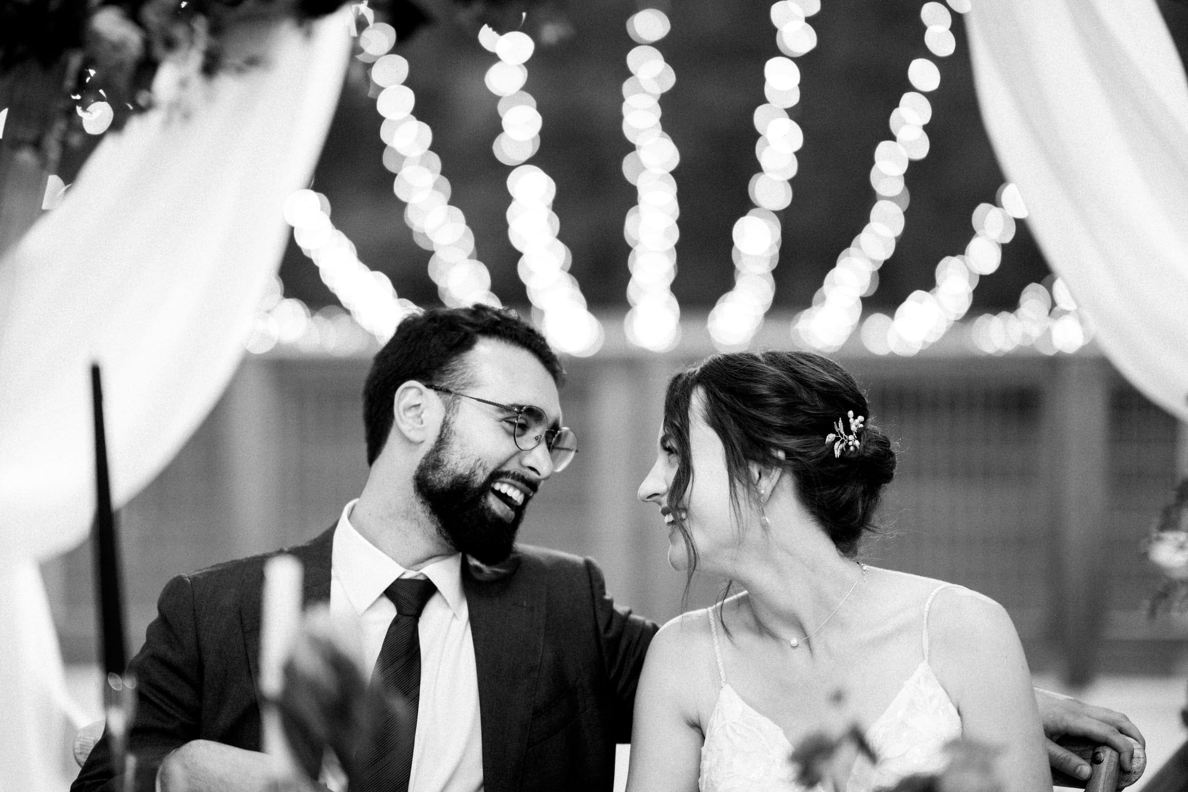 union grove farm wedding photography - kaylyn & jeremy A black and white image captures a couple seated closely, smiling at each other during their Union Grove Farm wedding. The man in a suit and glasses complements the woman’s light dress, while blurred lights and draped fabric create a festive backdrop, reflecting timeless love.