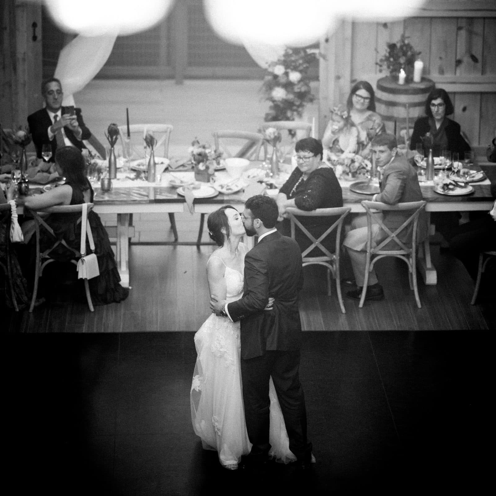 union grove farm wedding photography - kaylyn & jeremy A black and white photo captures a couple sharing their first dance at a Union Grove Farm wedding reception. Embraced on a dark dance floor, they're surrounded by seated guests capturing the moment at wooden tables adorned with flowers and candles.