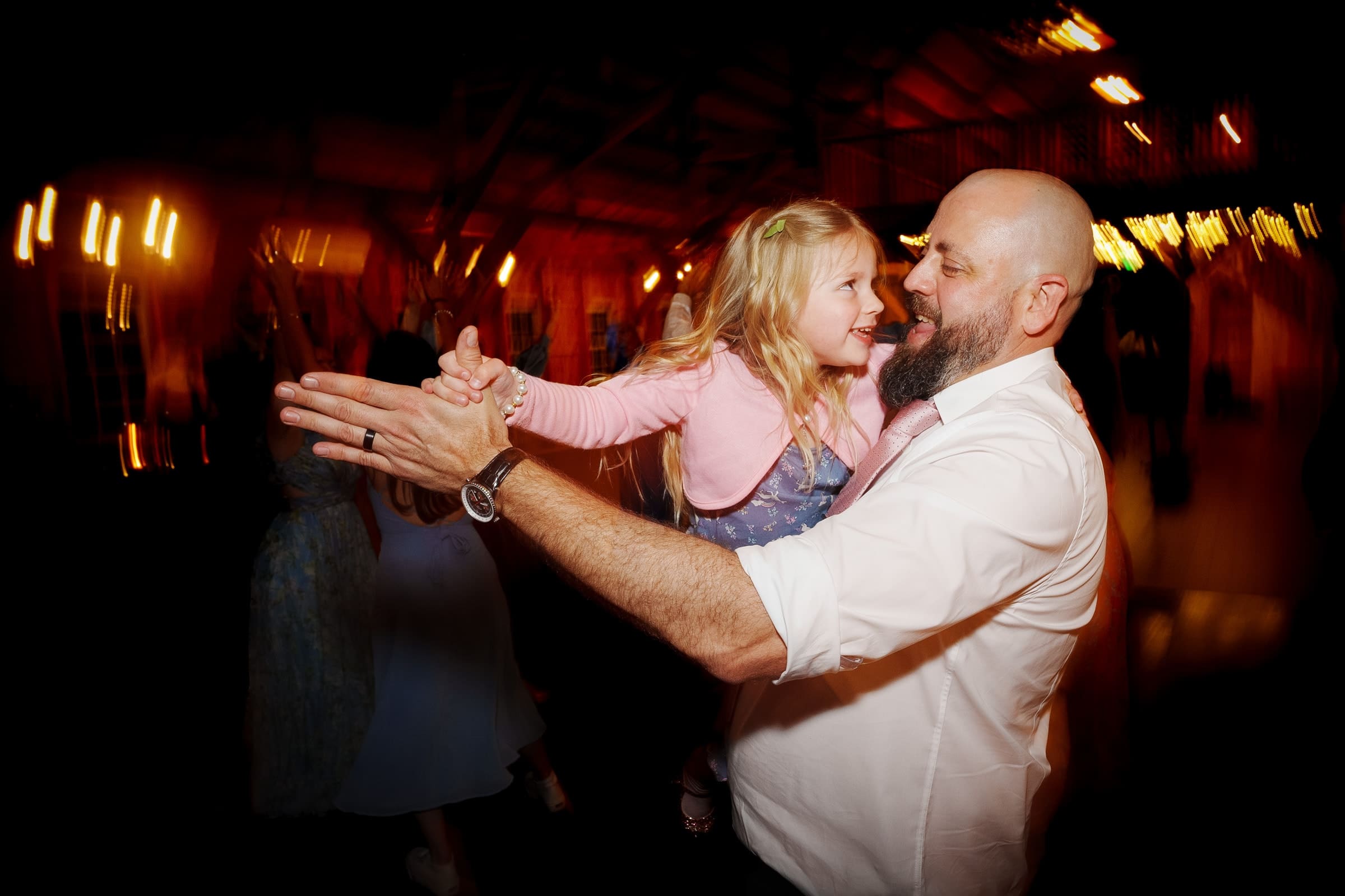 union grove farm wedding photography - kaylyn & jeremy A bearded man in a white shirt smiles as he dances with a laughing young girl in a pink dress at an indoor event, captured beautifully by Union Grove Farm Wedding Photography. The background is softly blurred with warm lighting and people mingling.