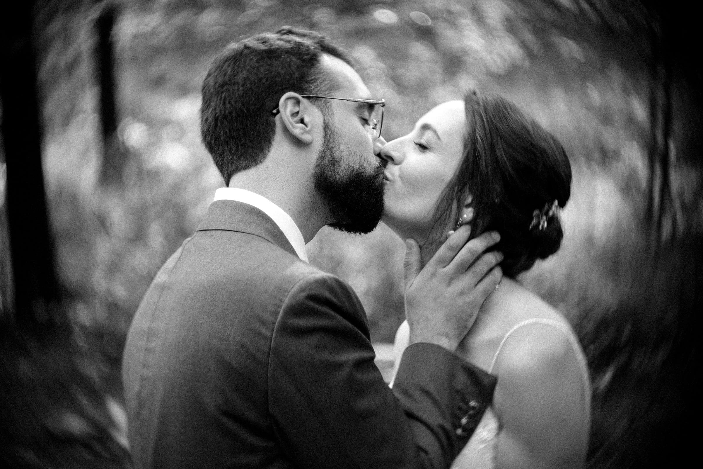 union grove farm wedding photography - kaylyn & jeremy A black and white photo of a couple kissing at Union Grove Farm. The man, with a beard and glasses, gently holds the woman's face. Her hair is styled up with a hairpiece, creating an intimate moment captured by wedding photography against a blurred natural background.