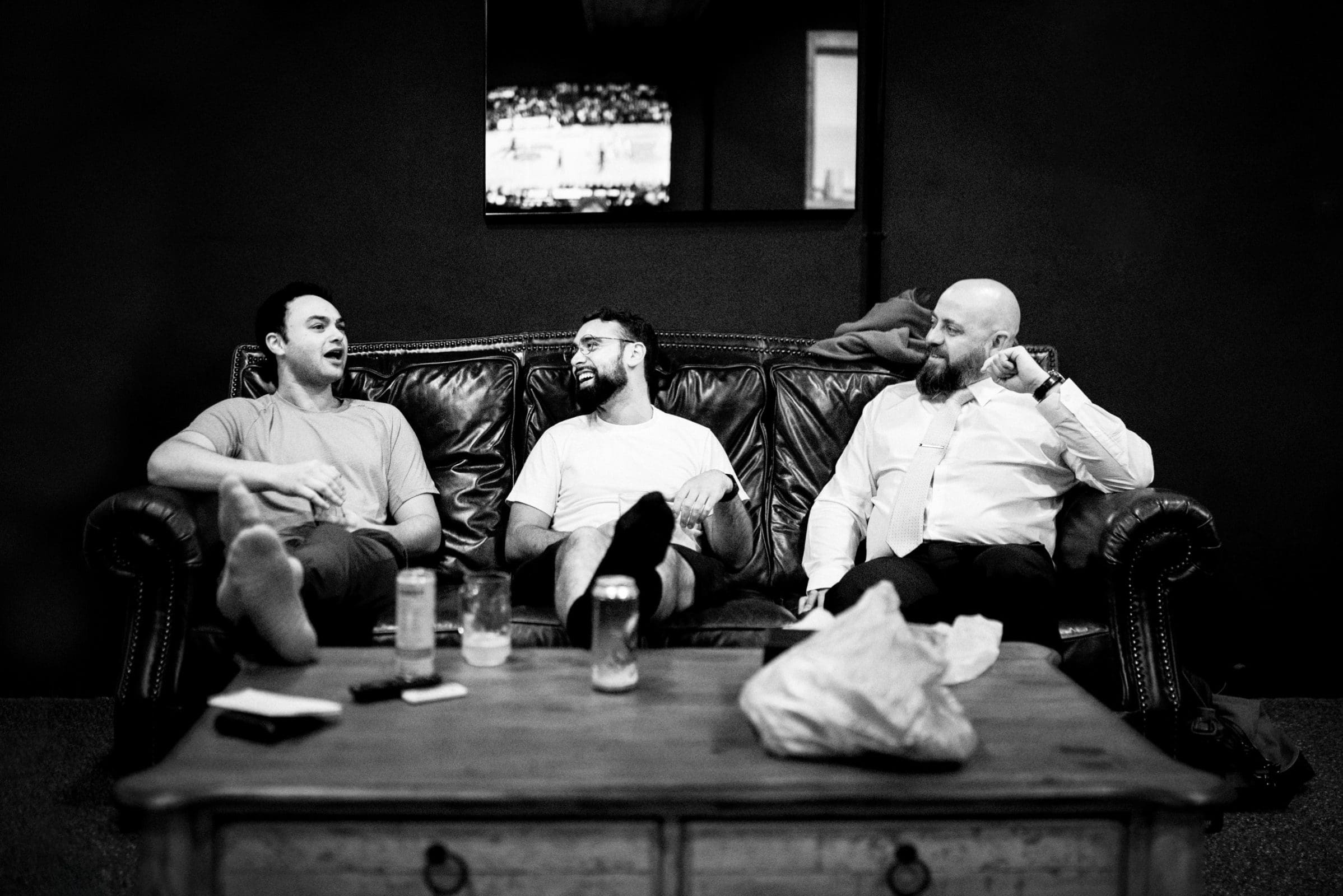 union grove farm wedding photography - kaylyn & jeremy Three men sit on a leather couch engaged in conversation, their camaraderie reminiscent of a cozy gathering at Union Grove Farm. The left man gestures animatedly, the middle man laughs heartily, and the right listens attentively. Drinks rest on the coffee table, while a TV subtly plays in the background.