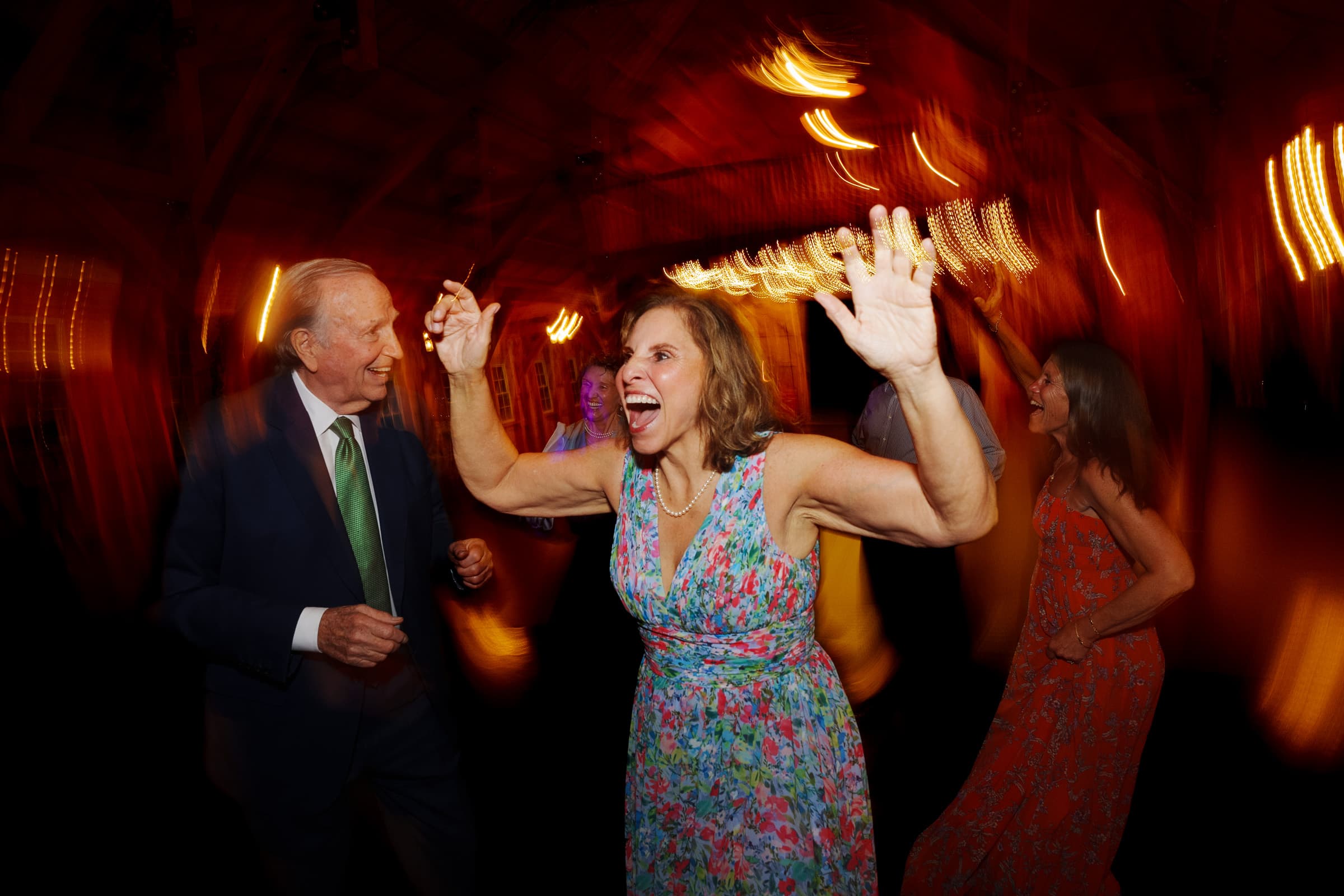 union grove farm wedding photography - kaylyn & jeremy People joyfully dancing in a warmly lit room at a Union Grove Farm celebration. A woman in a colorful floral dress has her arms raised, smiling exuberantly, while others join in. The motion blur of the wedding photography captures the energy and excitement of the joyous scene.
