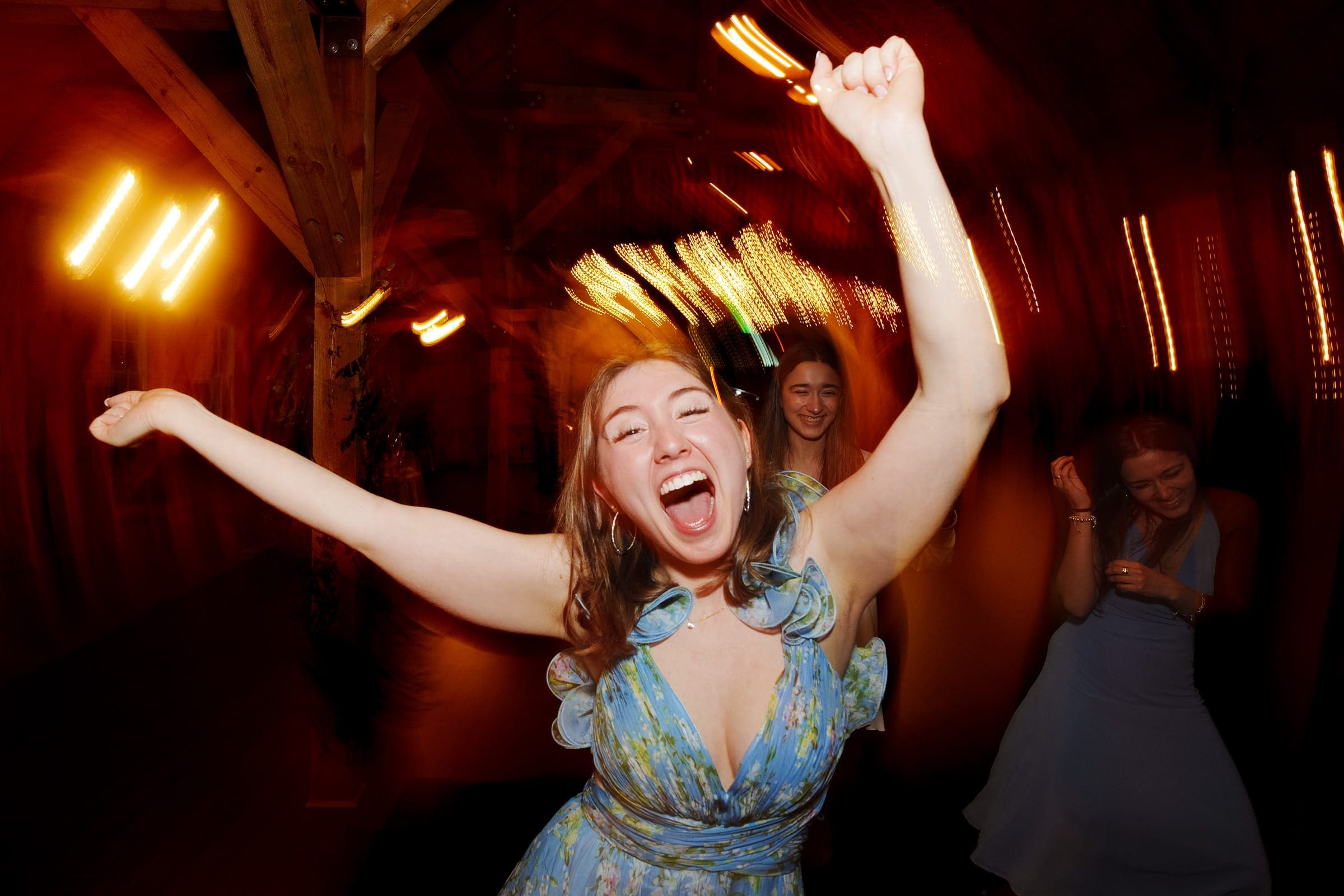 union grove farm wedding photography - kaylyn & jeremy A woman in a blue floral dress energetically dances at the Union Grove Farm wedding, her joyful expression matched by two others in similar dresses. With arms in the air and warm light trails creating a dynamic atmosphere, this moment of celebration is beautifully captured in the photography.