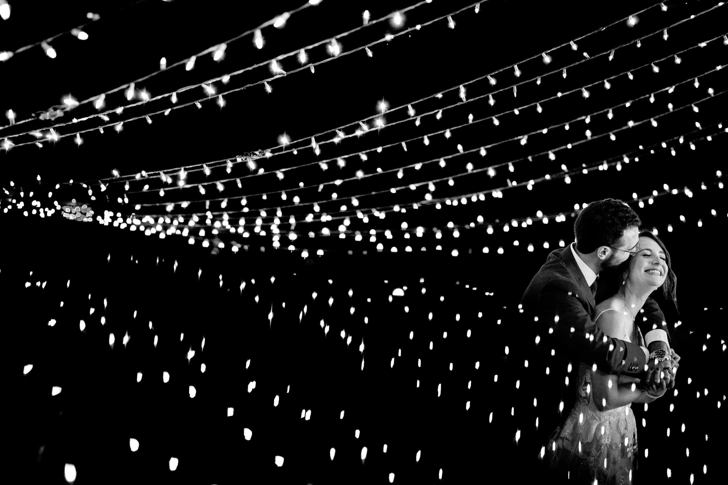 union grove farm wedding photography - kaylyn & jeremy A couple embraces under strings of twinkling lights at night. The black and white photograph captures their joyful moment at Union Grove Farm, surrounded by a canopy of glowing bulbs, creating a dreamy, romantic atmosphere perfect for wedding photography.