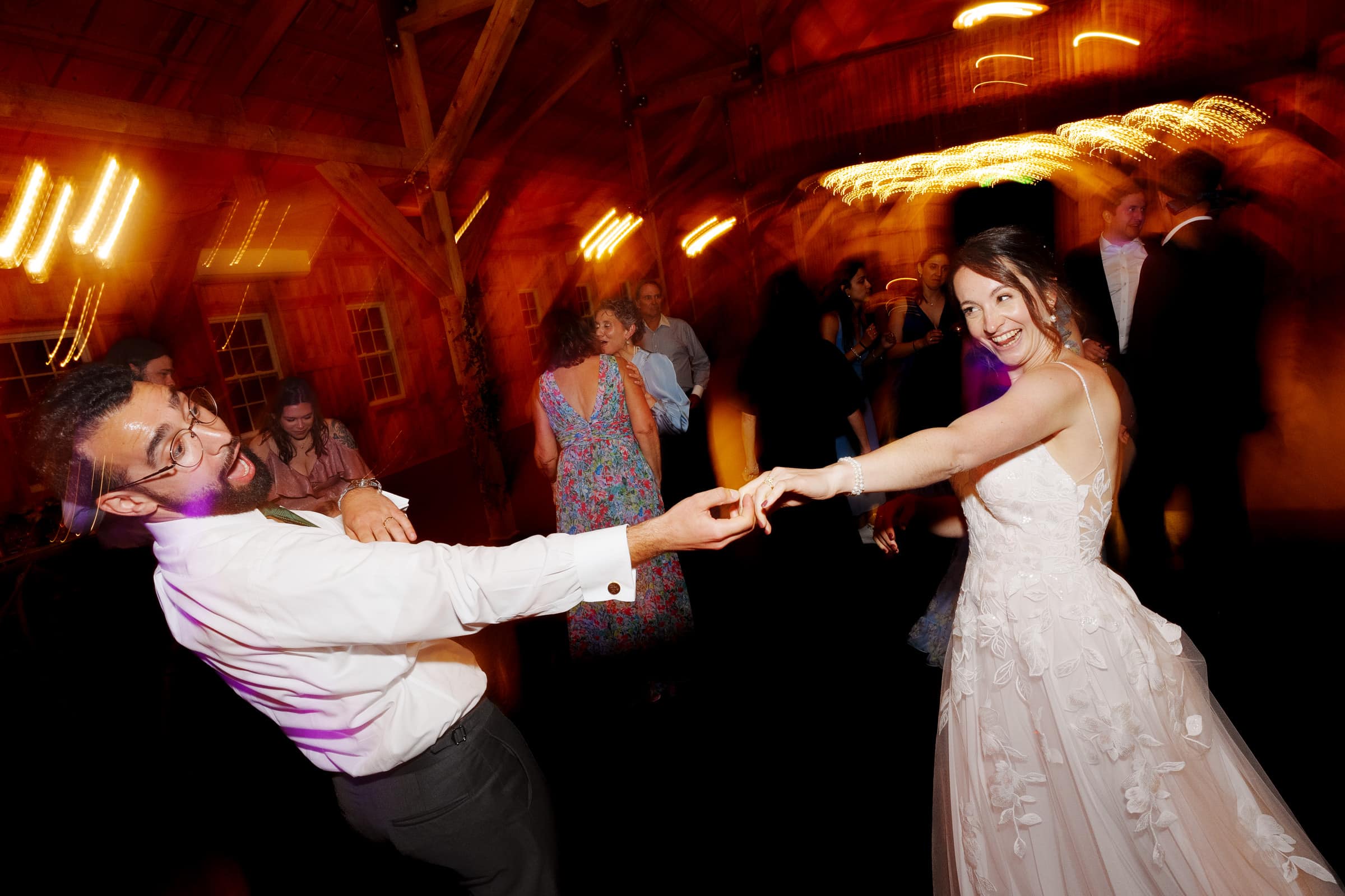 union grove farm wedding photography - kaylyn & jeremy A couple joyfully dances at an indoor union grove farm wedding. The man, wearing a shirt and tie, leans back with enthusiasm while the woman in her bridal gown smiles as they hold hands. The background captures other guests and the warm, blurred lights of a perfect celebration.