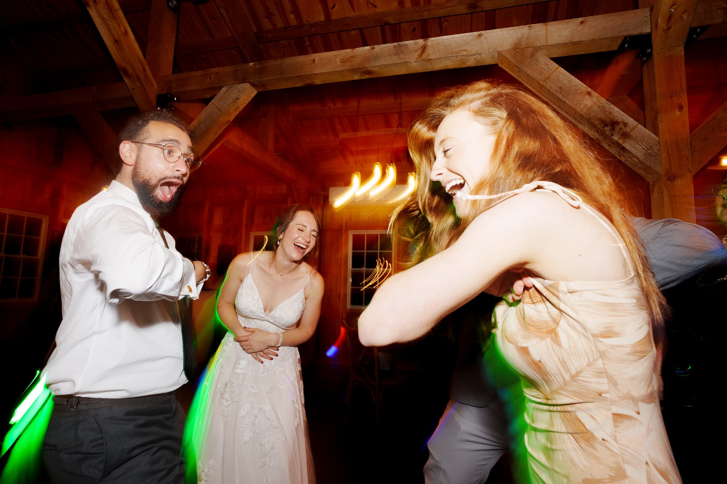 union grove farm wedding photography - kaylyn & jeremy A group of people joyfully dancing at a wedding reception, captured in the enchanting ambiance of Union Grove Farm. A man and two women, one in a white dress, are mid-dance in the warmly lit wooden venue, with colorful light trails elevating the lively atmosphere.