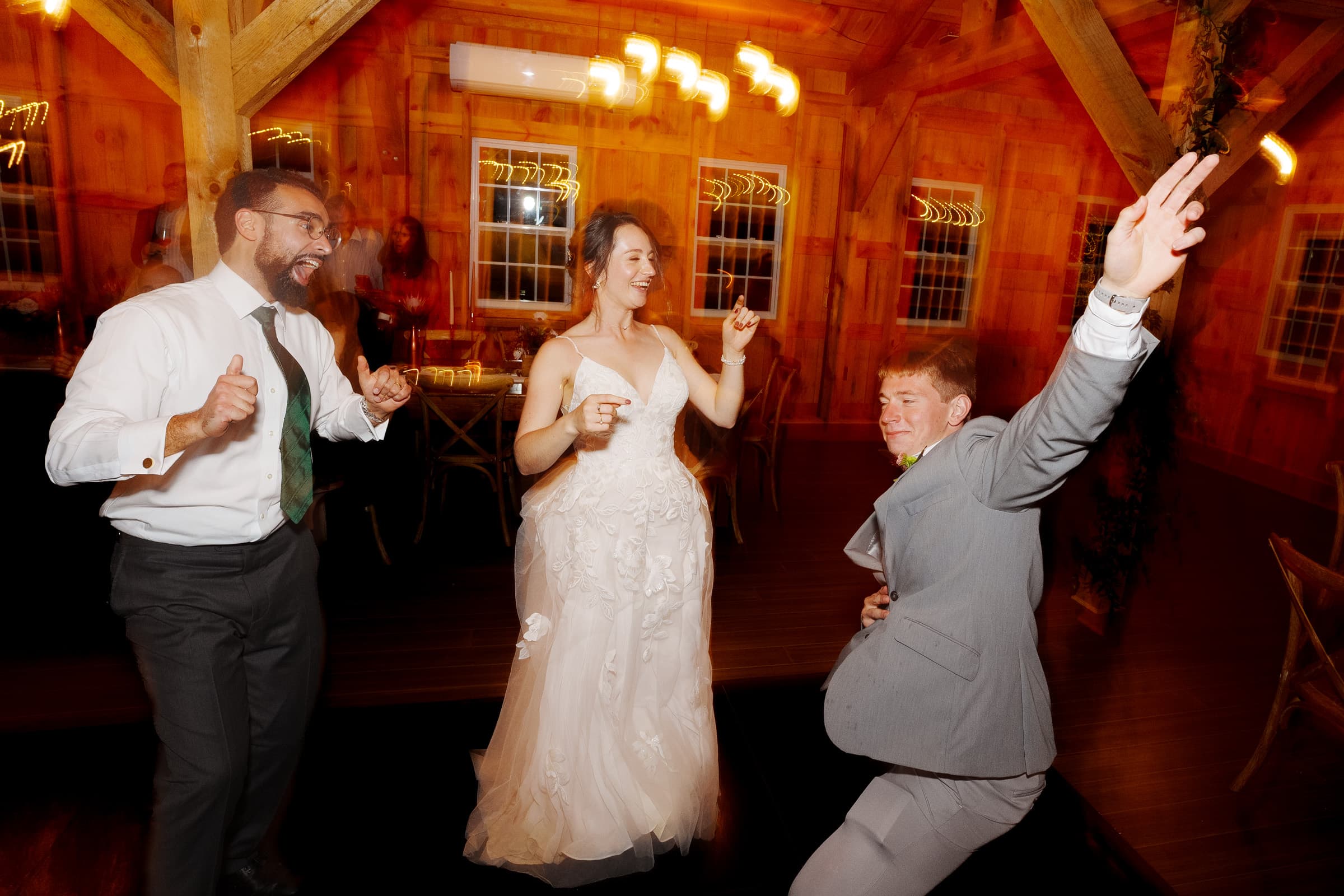 union grove farm wedding photography - kaylyn & jeremy Three people are dancing energetically in a warmly lit wooden room at a Union Grove Farm wedding. A woman in a white dress is smiling in the center, flanked by two men in suits, all enjoying the moment. The lively atmosphere exudes joy, perfectly captured by skilled photography.