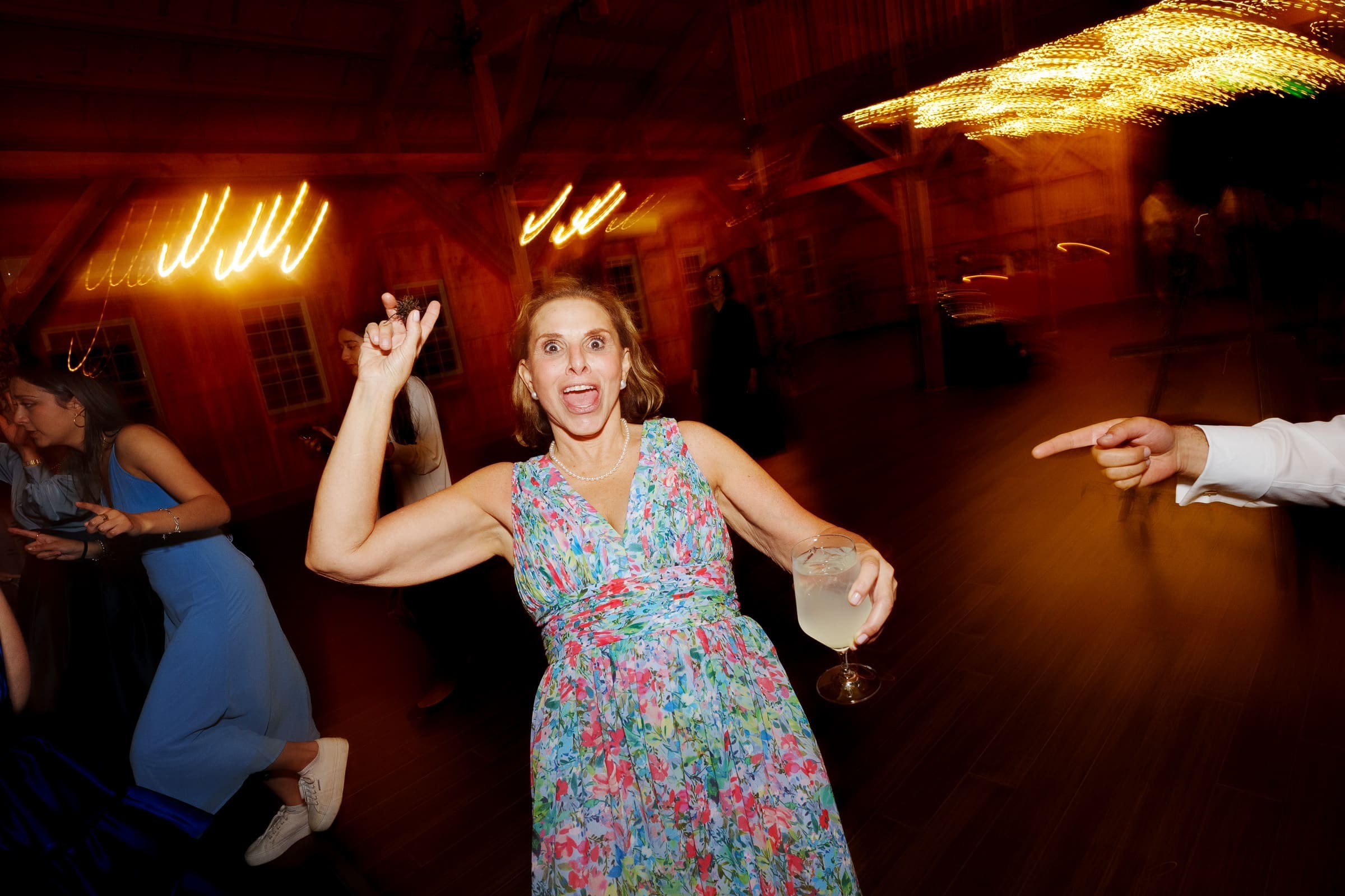 union grove farm wedding photography - kaylyn & jeremy A woman in a floral dress dances energetically at a union grove farm wedding, holding a drink in one hand. Motion blur and warm lighting enhance the lively atmosphere. Another hand points towards her from the right side of the image, capturing the vibrant essence of celebration and photography.