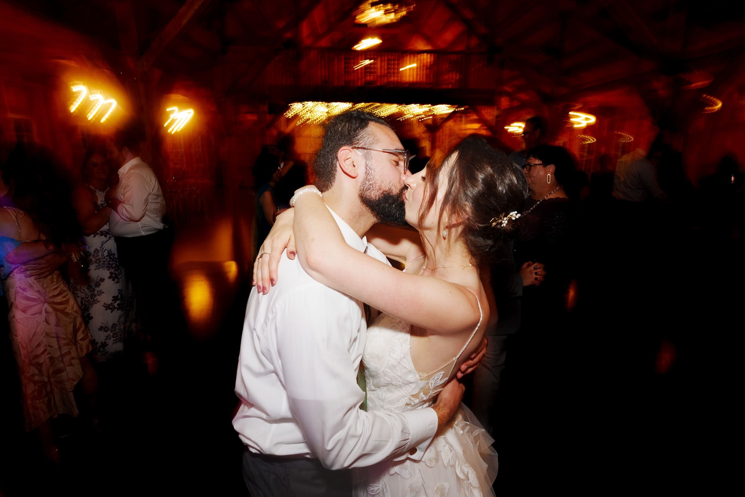 union grove farm wedding photography - kaylyn & jeremy A couple shares a romantic kiss on the dance floor at Union Grove Farm. The dimly lit venue, captured through wedding photography, features blurred lights and guests dancing in the background, creating a warm and intimate atmosphere.