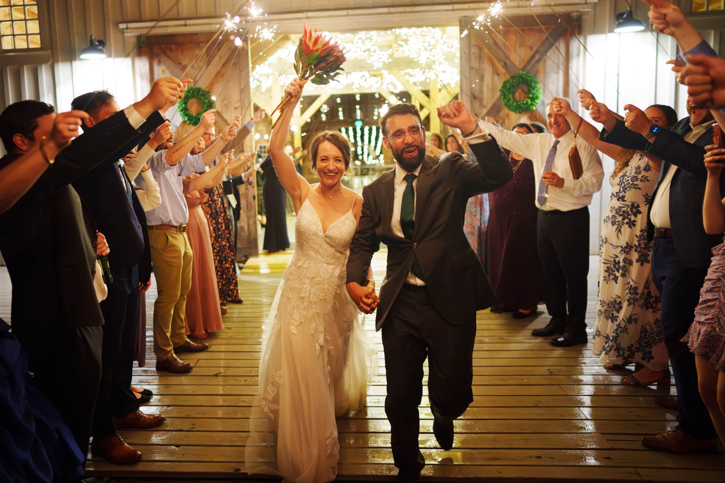 union grove farm wedding photography - kaylyn & jeremy A joyful bride and groom walk hand in hand under a sparklers arch at Union Grove Farm, surrounded by smiling guests. The bride holds a bouquet, and both wear formal attire. The warmly lit venue provides a festive backdrop, perfect for capturing this beautiful wedding photography moment.