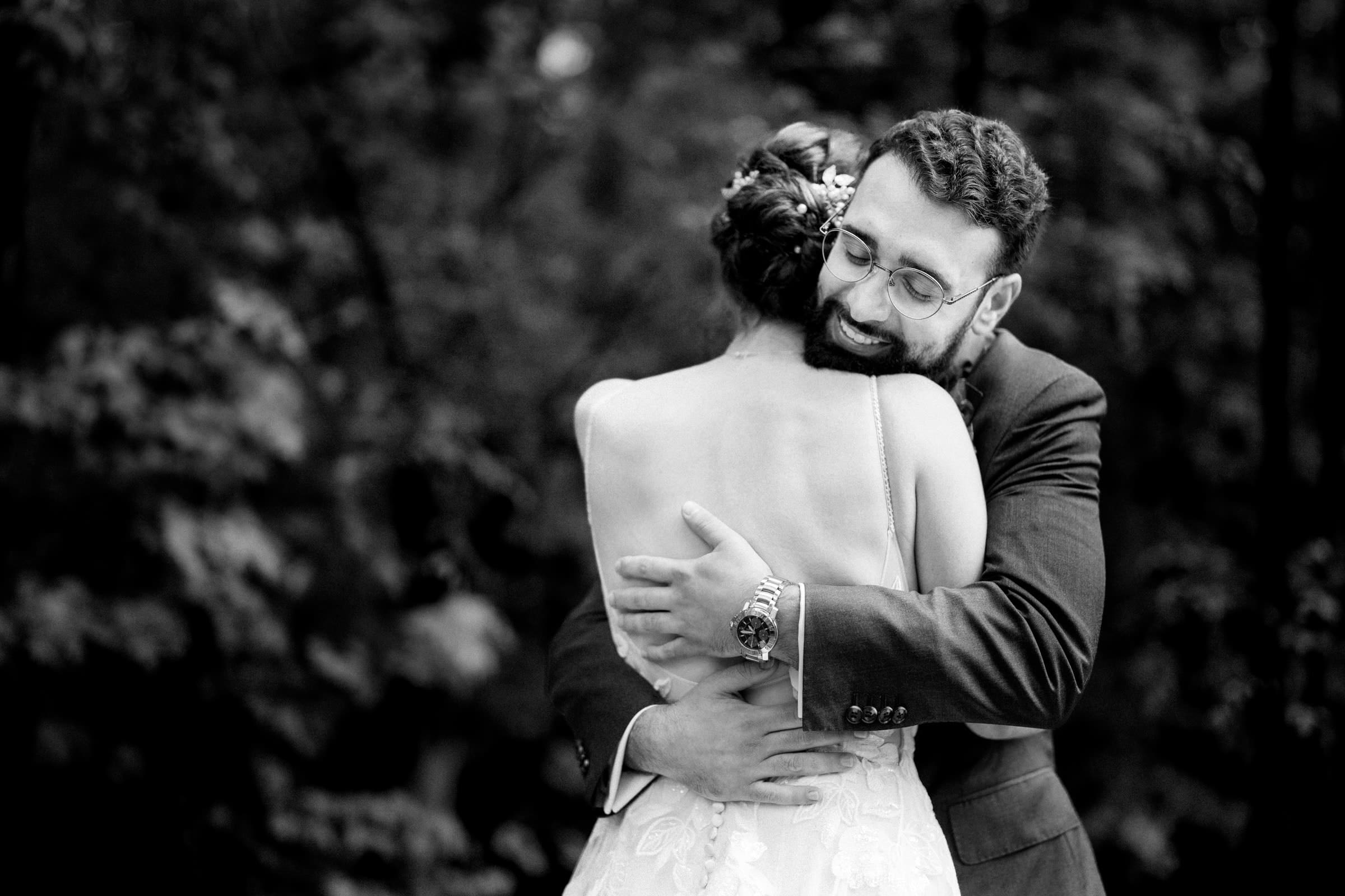 union grove farm wedding photography - kaylyn & jeremy A black-and-white image captures a couple embracing at Union Grove Farm. The man, donning glasses and a beard in a suit, lovingly hugs the woman in a strapless dress with floral details. They stand outdoors, with blurred foliage adding depth to this beautiful wedding photography moment.