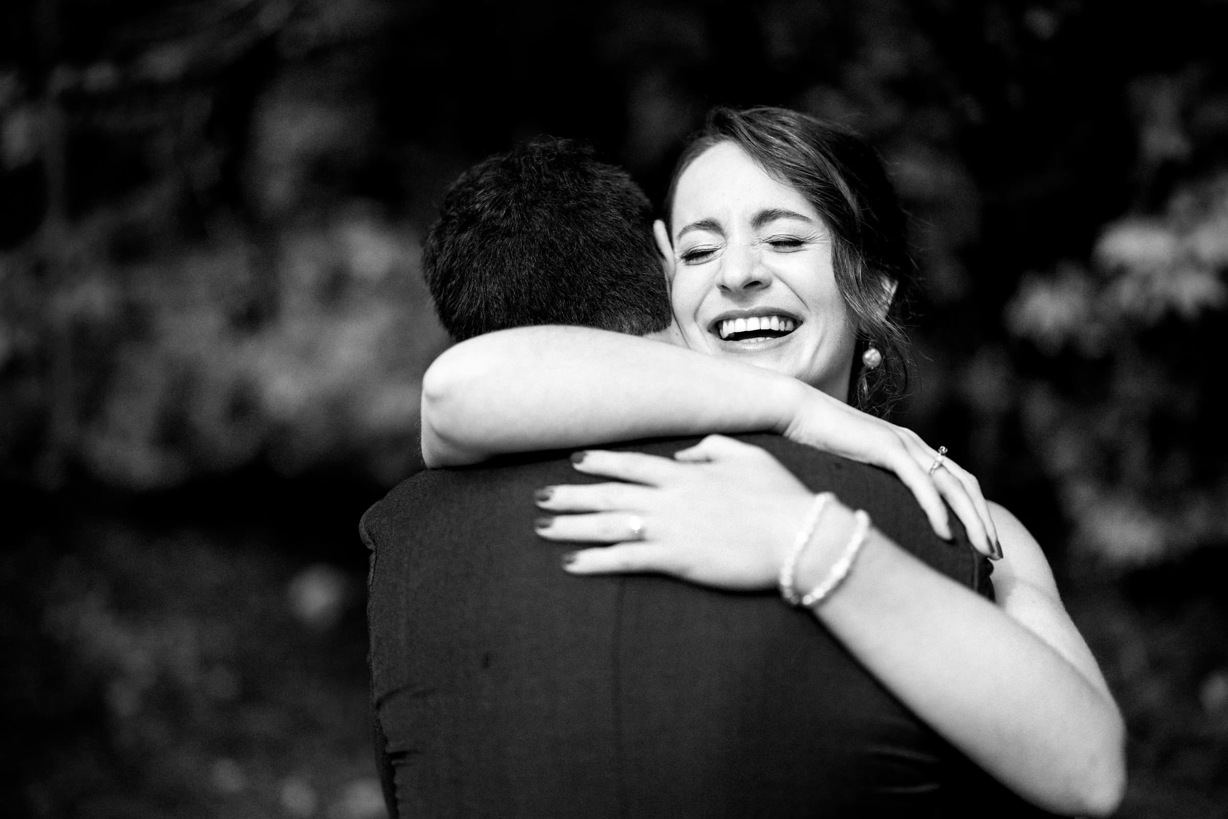 union grove farm wedding photography - kaylyn & jeremy A woman joyfully embraces a man in a black-and-white setting, capturing the essence of union at Grove Farm. With her wide smile and closed eyes, she embodies pure bliss. Her bracelet glints as she holds him tight, his back to the camera against a backdrop softly blurred with trees.