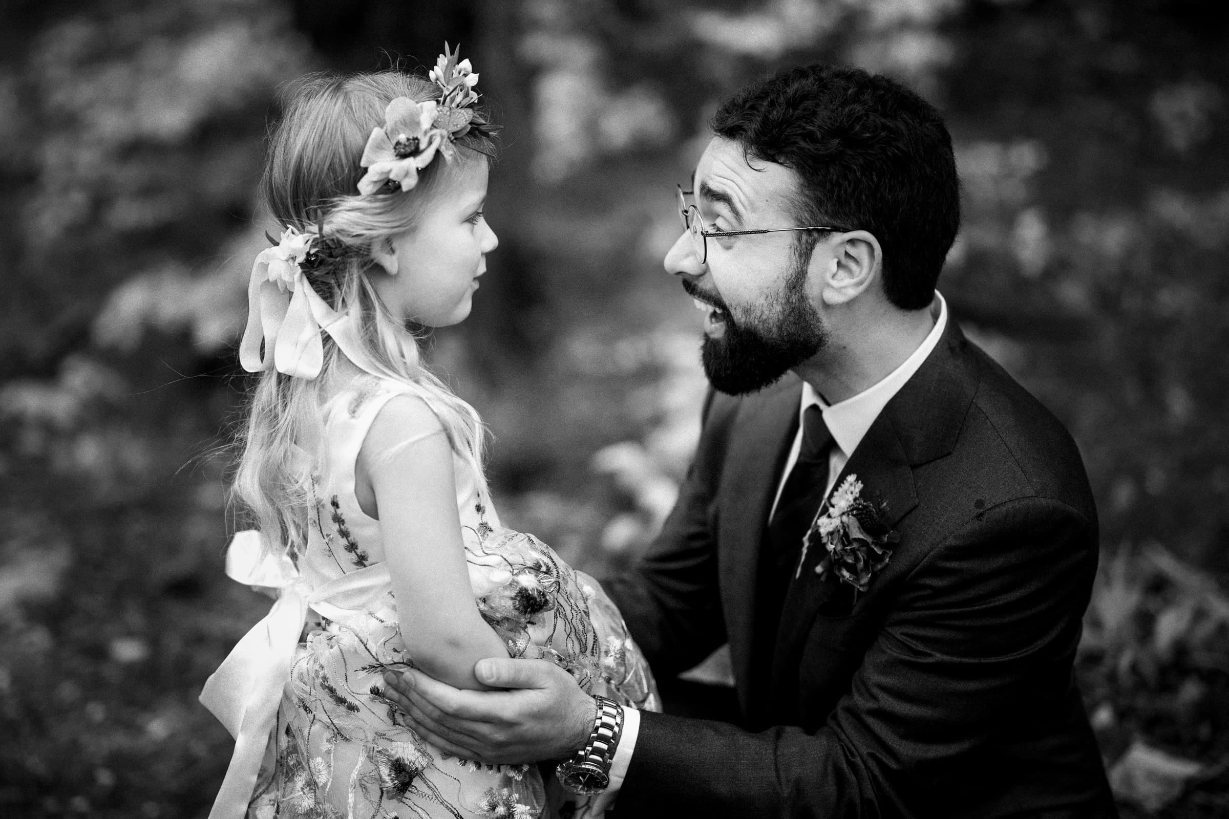 union grove farm wedding photography - kaylyn & jeremy In a timeless black and white photo, the groom shares a joyful moment with a young girl in a flower dress and floral headband. Set amid the lush surroundings of Union Grove Farm, his warm smile captures the essence of wedding photography at its finest.