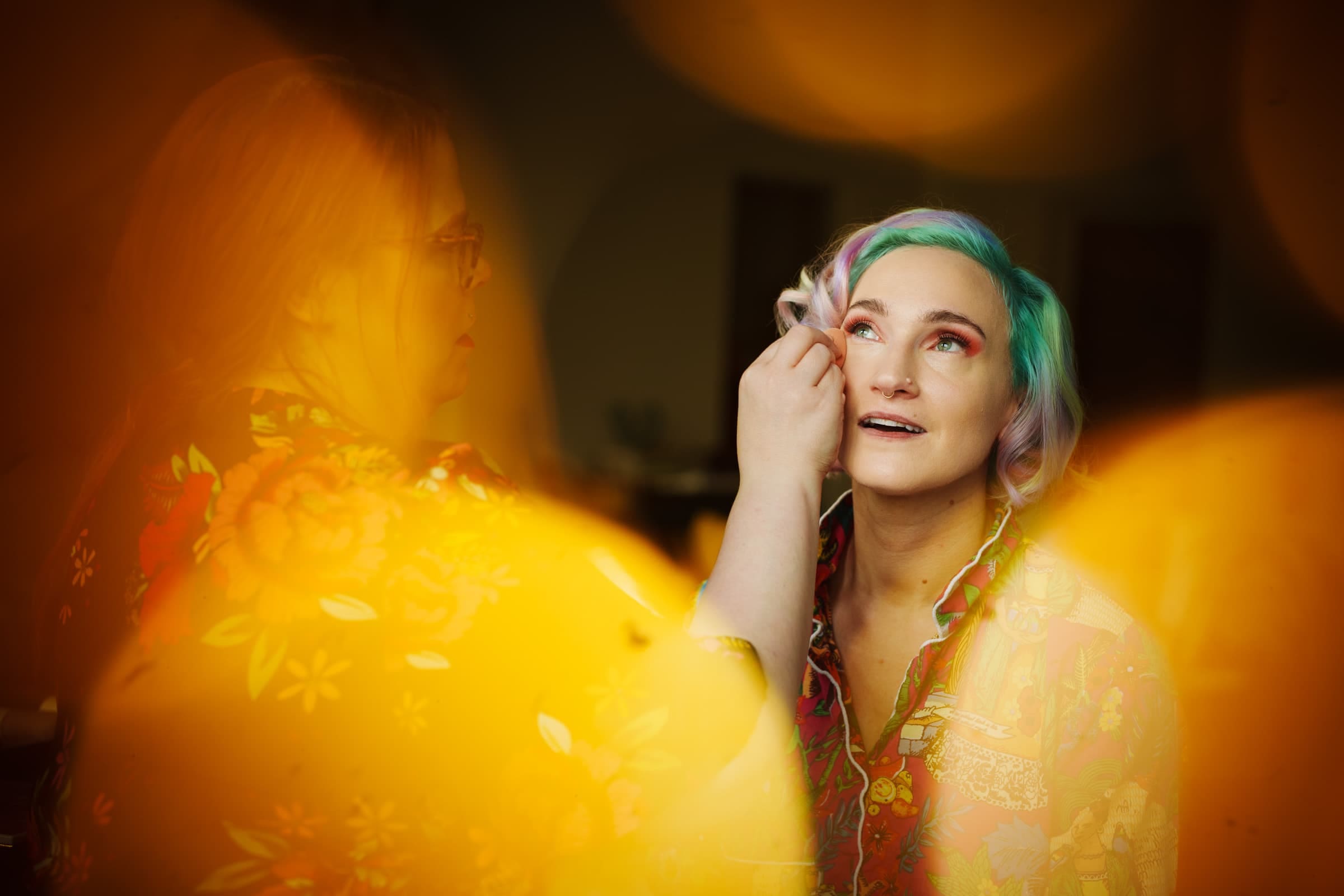 hideaway asheville wedding photography - payne & phil A person with colorful hair and vibrant makeup is being assisted by another, surrounded by warm, diffused light, creating a dreamy atmosphere. Both individuals in vivid attire add to the artistic scene, reminiscent of a Hideaway Asheville wedding photography session.