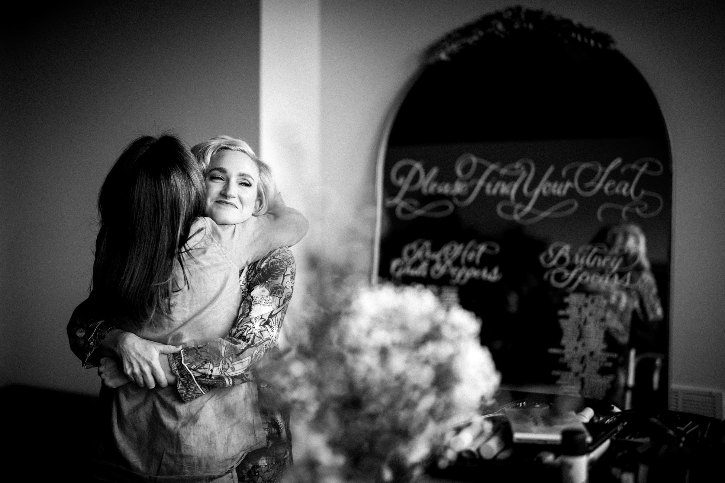 hideaway asheville wedding photography - payne & phil A black and white photo captures two people embracing warmly in front of a mirror with calligraphy text, evoking the intimate charm of a hideaway. One person, smiling with short hair, holds the other who has long hair facing away. A blurred bouquet in the foreground adds a dash of Asheville wedding photography allure.