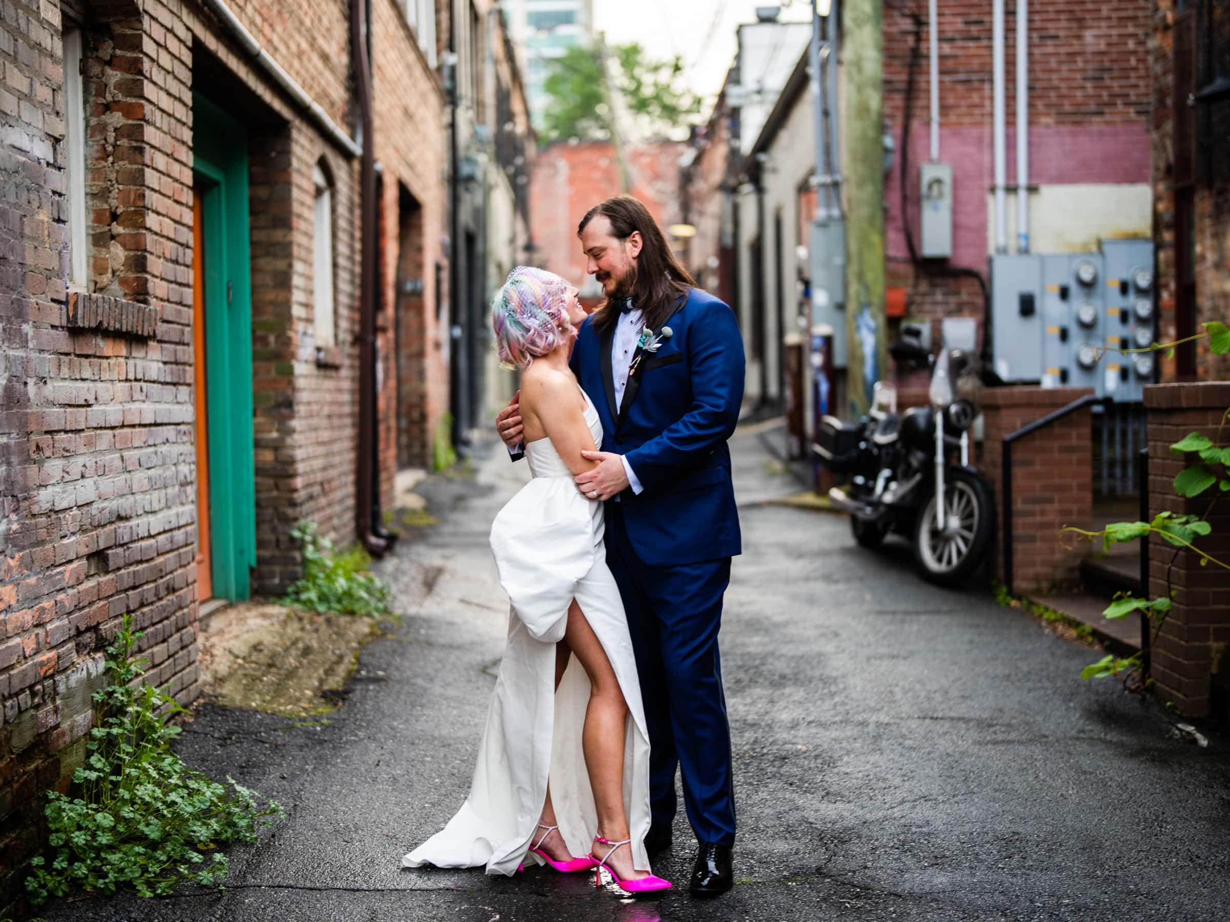 A couple embraces in a narrow alleyway, with the man in a dark blue suit and the woman in a white dress and pink shoes, her colorful hair adding flair. A motorcycle and urban brick buildings peek from behind, capturing the essence of Hideaway Asheville wedding photography.