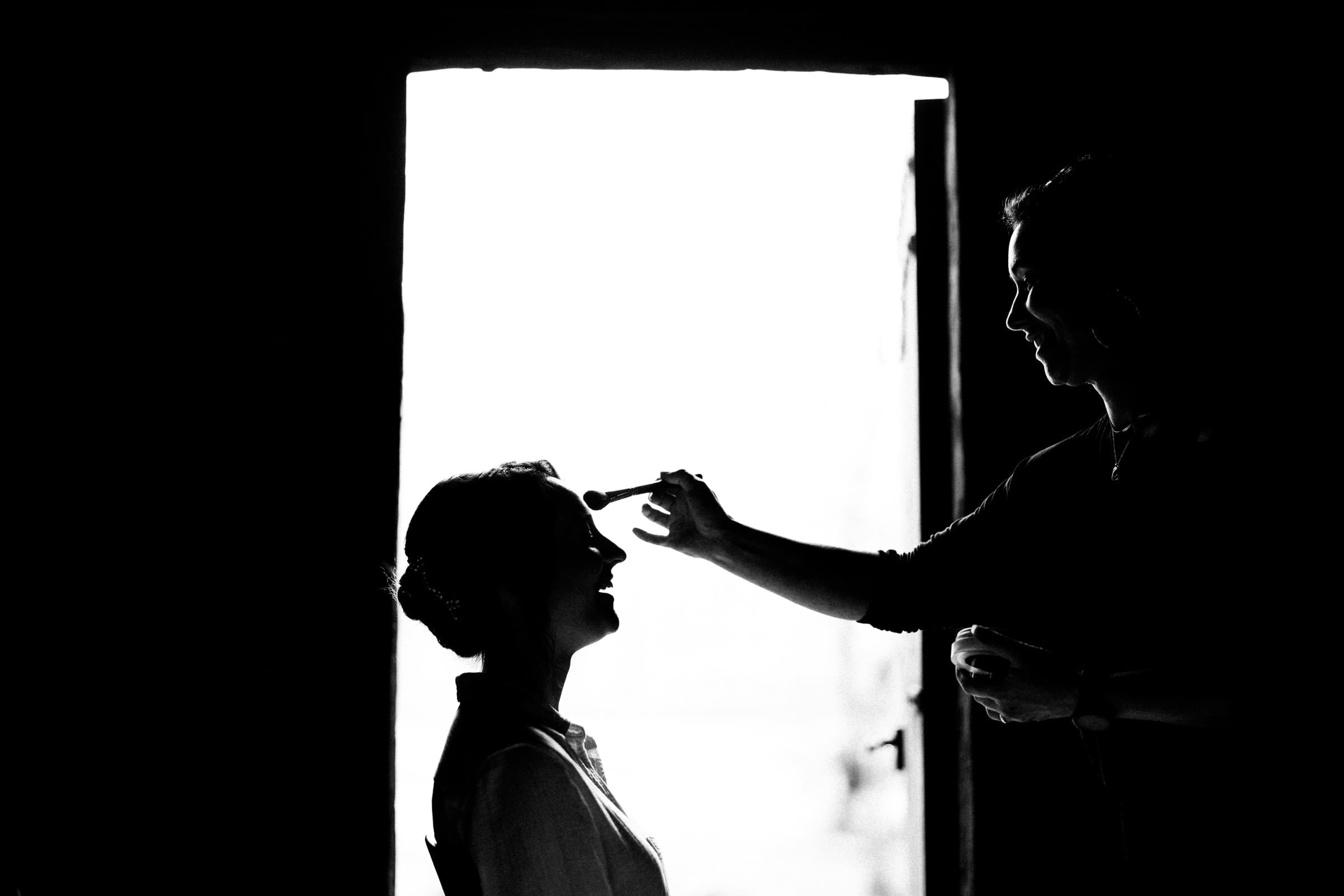 union grove farm wedding photography - kaylyn & jeremy A silhouette of a person applying makeup to another’s face captures the essence of serene preparation, reminiscent of Union Grove Farm's intimate wedding photography. The scene is backlit, with light streaming through an open door, highlighting their profiles in a dark room.