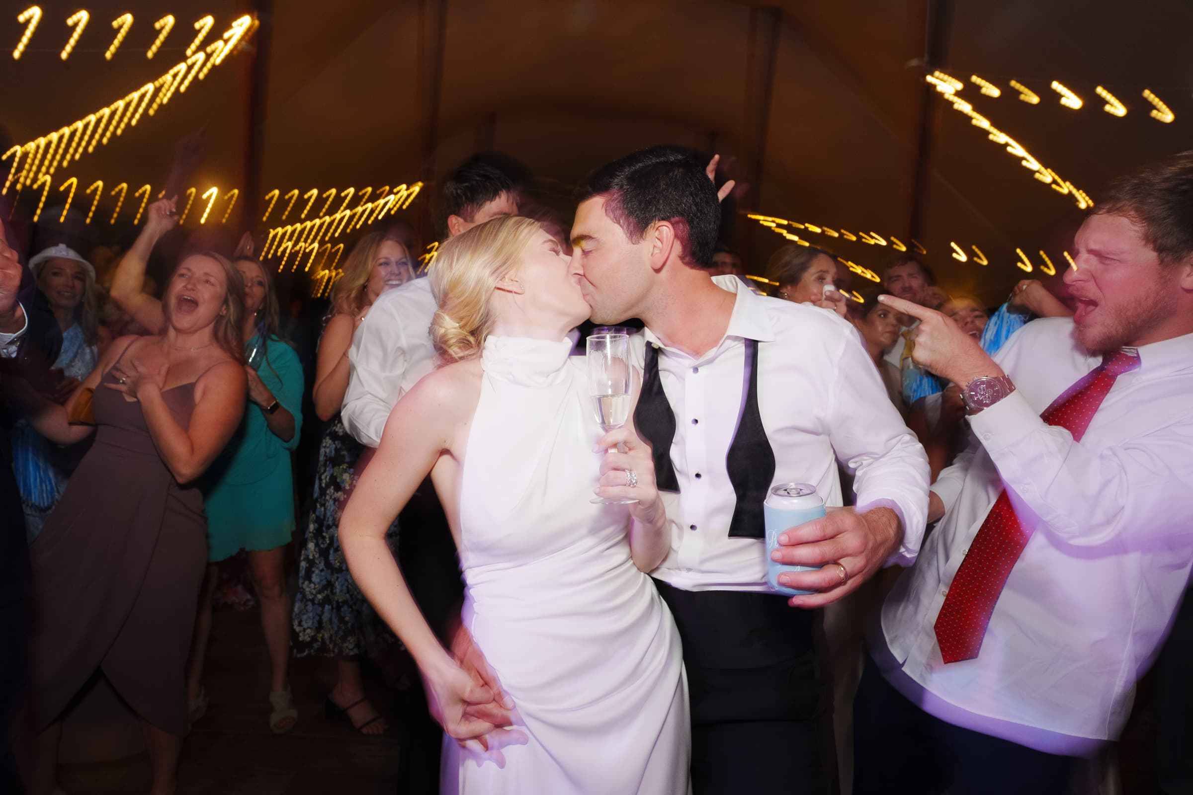 dunn wedding photography - reaves & zach A bride and groom share a kiss on the dance floor, captured perfectly by Dunn wedding photography, surrounded by cheering guests. The bride holds a champagne flute, while the groom has his arm around her. Festive lights and blurred motion create a lively atmosphere.