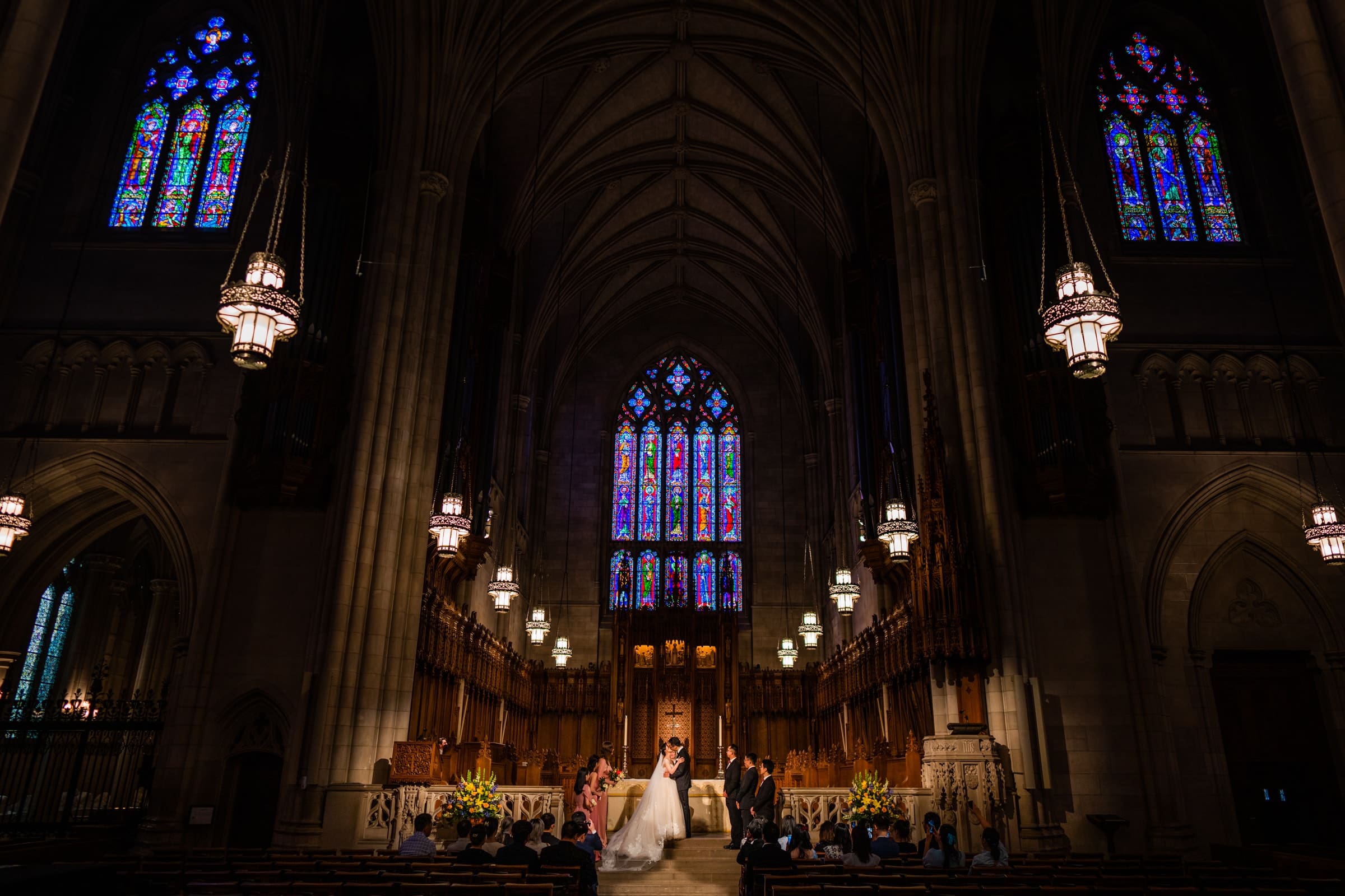 A wide-angle wedding photograph inside Duke Chapel during the ceremony, looking toward the altar under towering vaulted ceilings with three massive, colorful gothic stained-glass windows visible in the background.