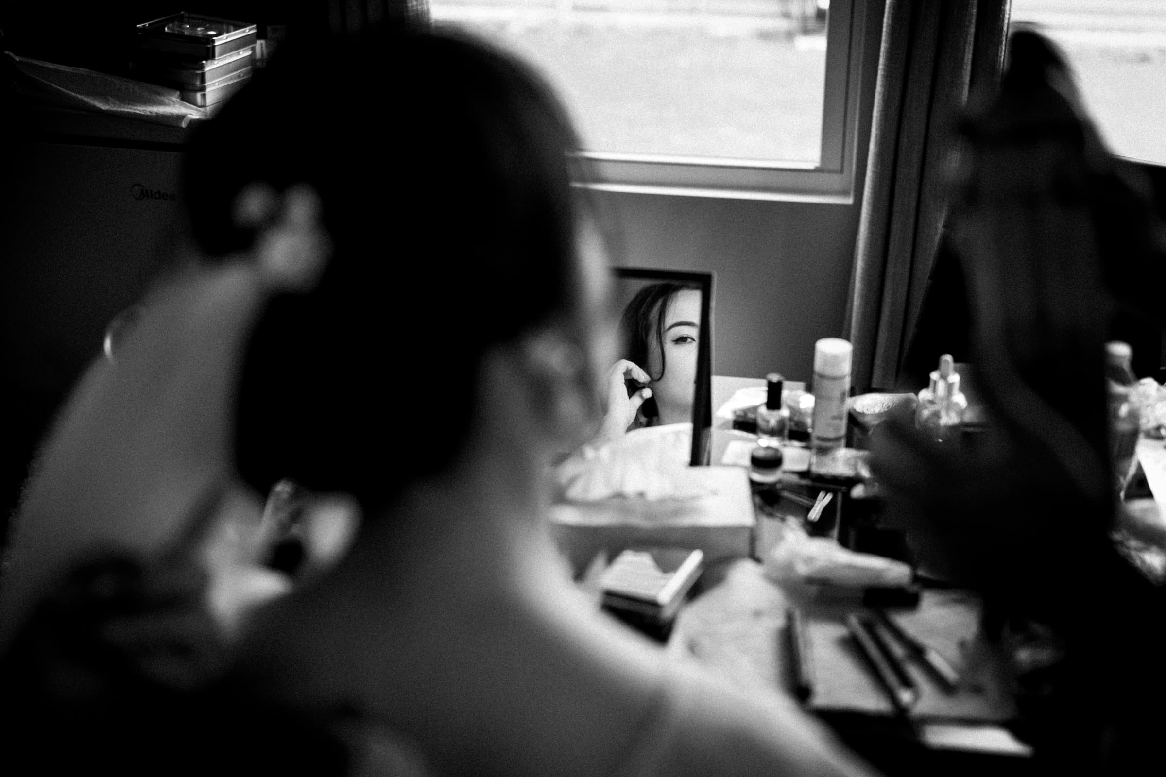 intimate duke chapel wedding - jenny & andrew A woman with her hair up is getting ready, gazing into a small mirror on a cluttered table of makeup products. The black and white image exudes a classic, timeless feel, reminiscent of preparations for an intimate Duke Chapel wedding.