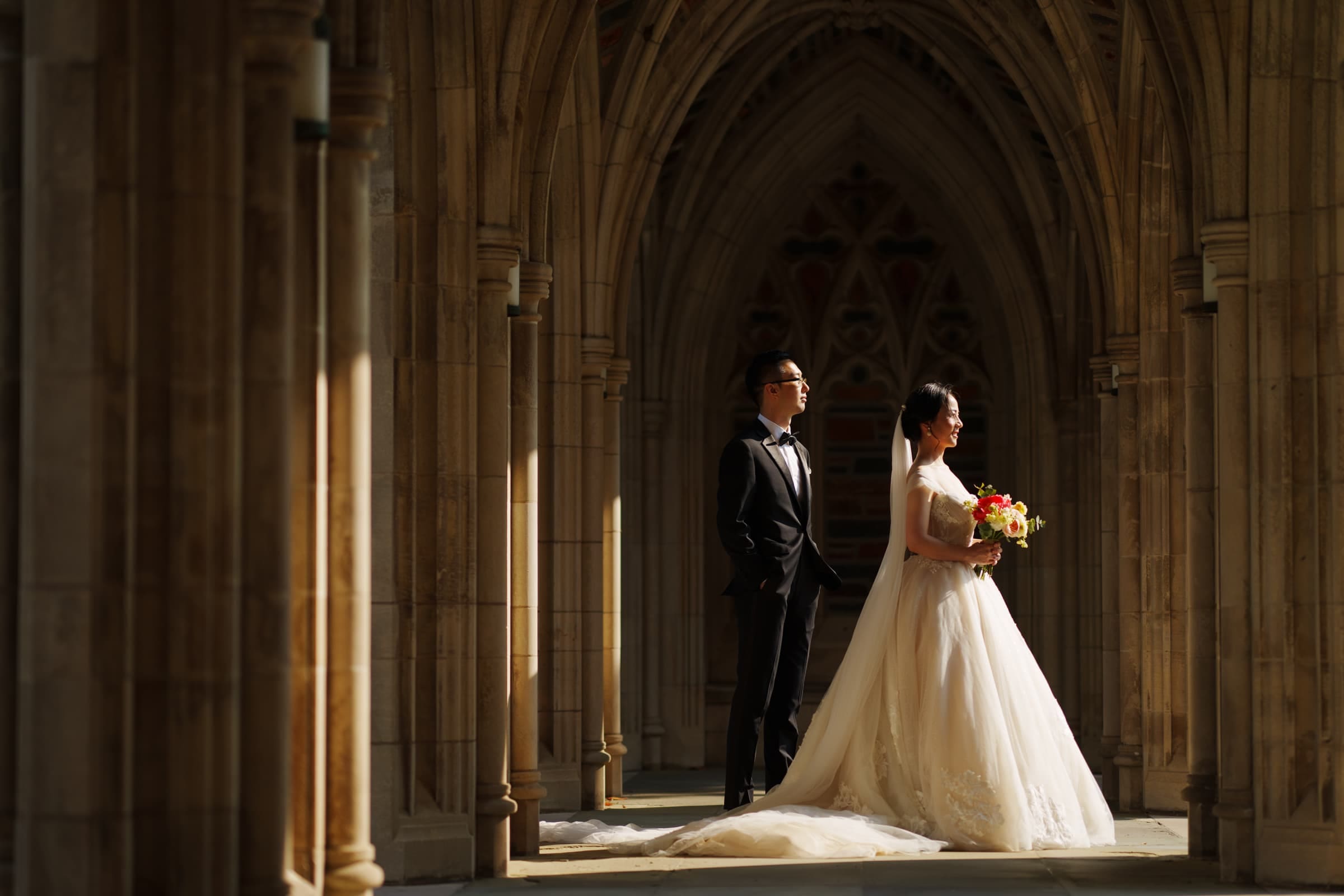 A couple in wedding attire stands under an ornate, sunlit stone archway at their intimate Duke Chapel wedding. The bride holds a bouquet of flowers, and both gaze forward, creating a serene and elegant scene.