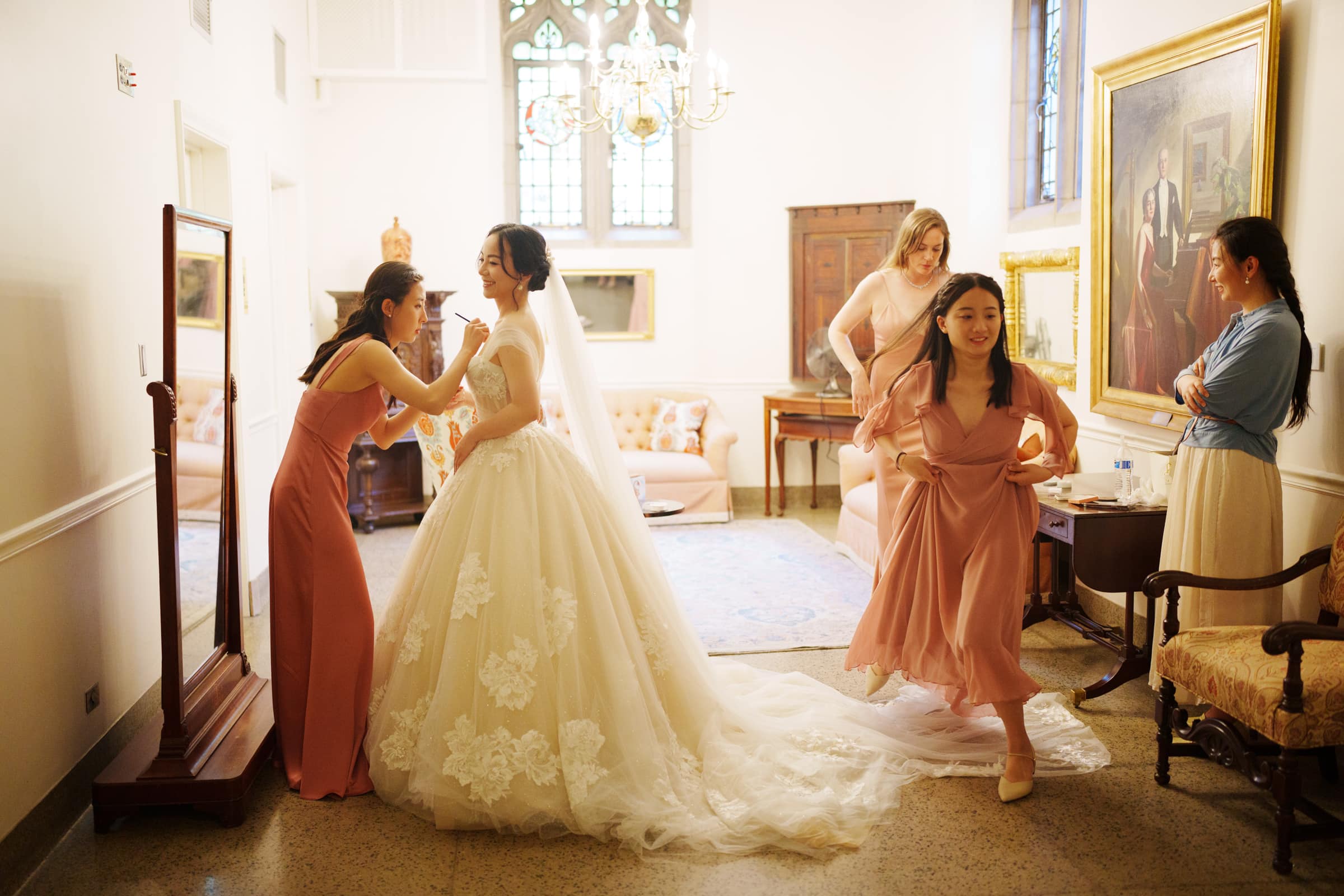 A bride in a white wedding gown is being assisted by two bridesmaids in a well-lit room, capturing an intimate moment reminiscent of a Duke Chapel wedding. Two other women are adjusting their dresses, while a woman stands nearby. The room boasts elegant furniture and paintings on the walls.