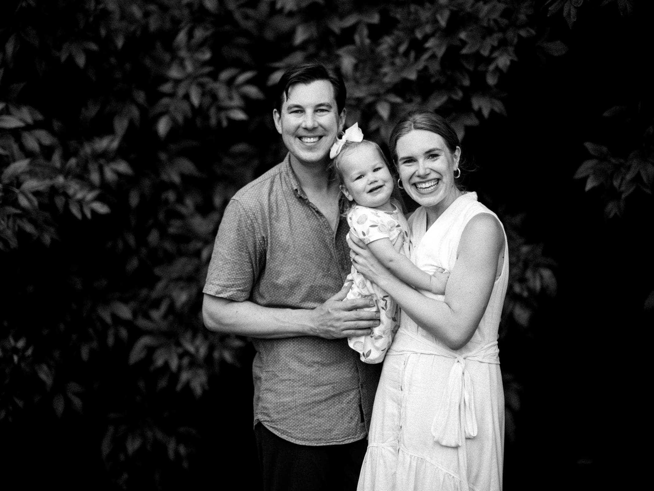 A timeless black and white family photography portrait captures a smiling group standing together. The man in a short-sleeve shirt holds a toddler adorned in a floral outfit with a bow, while the woman in a sleeveless dress stands beside them, all framed by leafy foliage in the background.