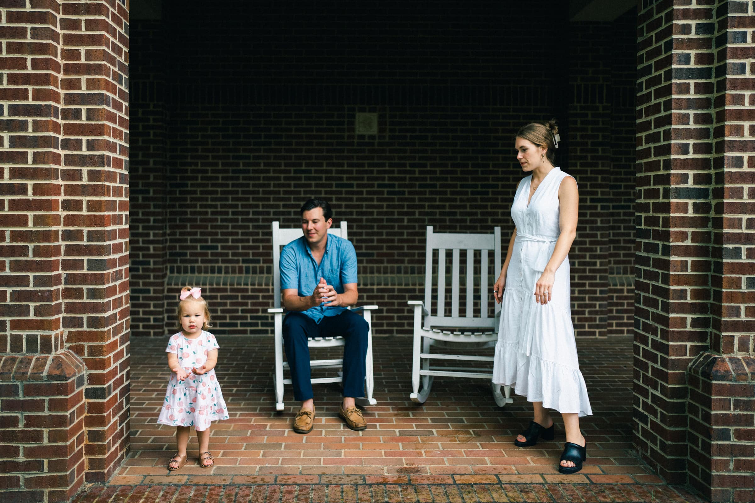 A toddler in a pink dress stands in front of two adults on white rocking chairs under a brick archway, capturing the essence of family photography. A woman in a white dress stands nearby, watching the child. The scene is relaxed and informal.