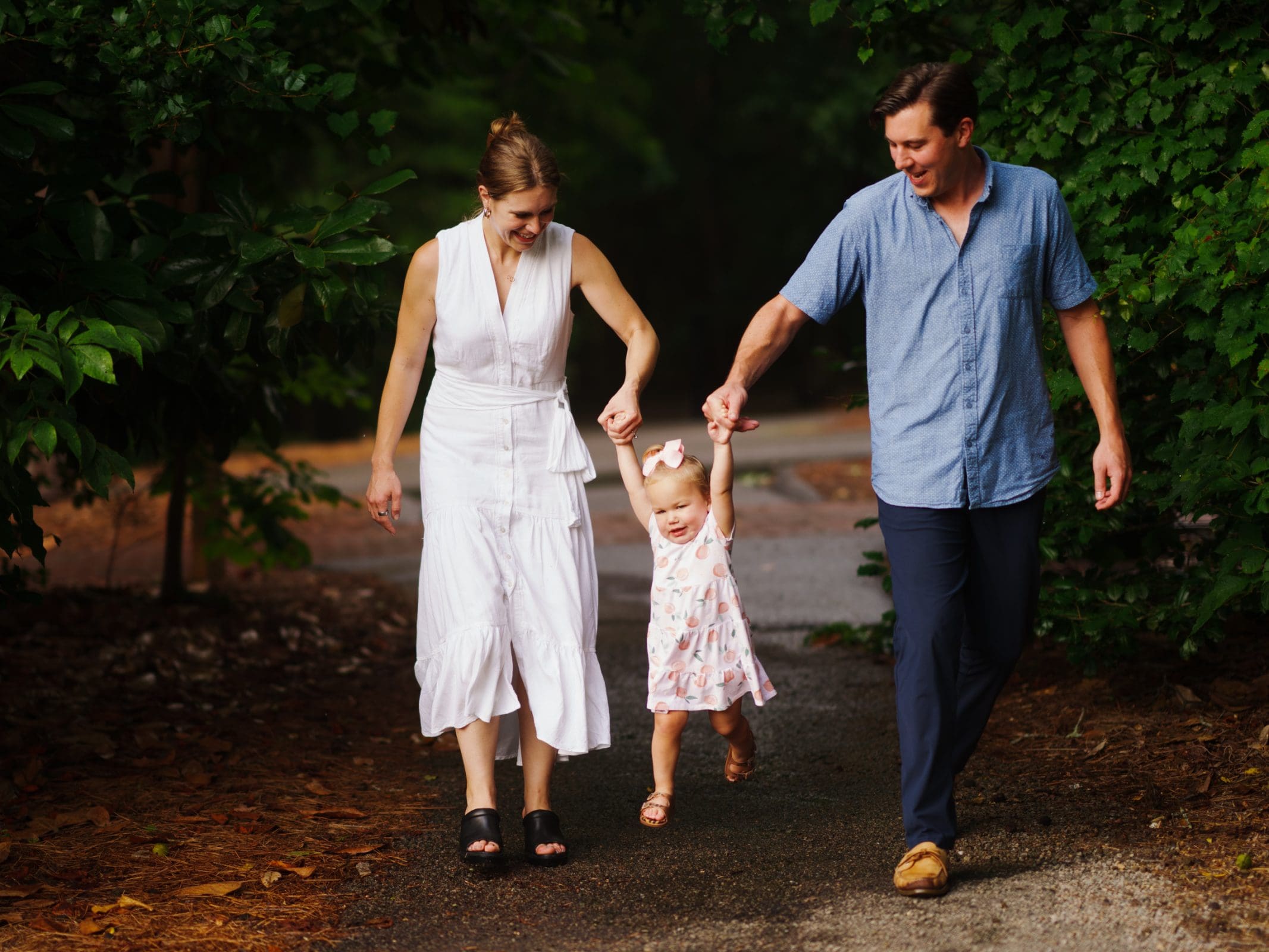 A family of three walks along a path surrounded by greenery. The mother in a white dress and the father in a blue shirt hold their young child’s hands, lifting her playfully between them. It's a perfect moment for family photography, capturing laughter and love in one frame.
