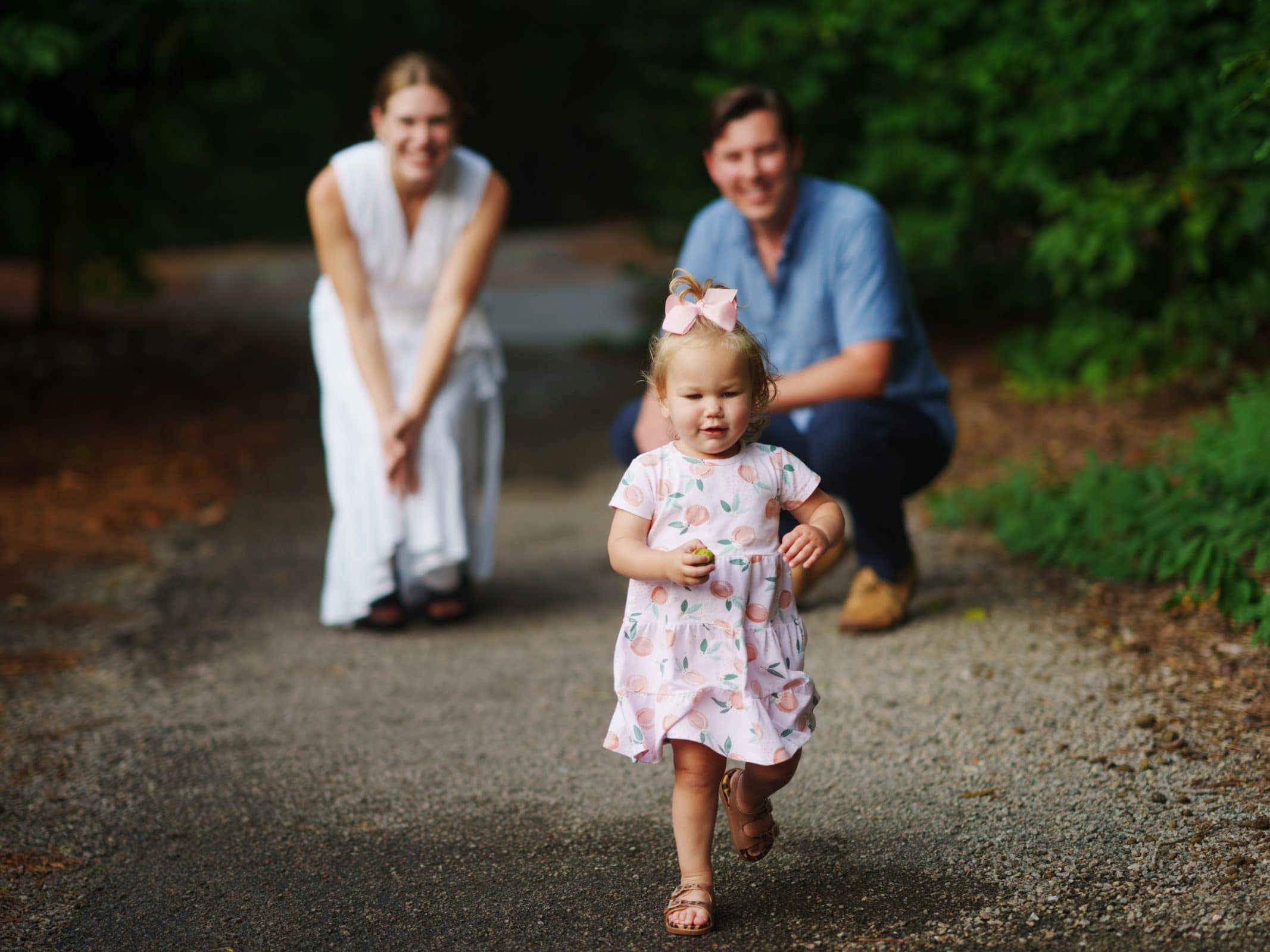 A toddler in a floral dress walks along a path, focused and holding a small object. In the background, two adults crouch down, smiling and watching her brisk adventure. The outdoor setting, surrounded by lush greenery, captures an authentic moment of family photography.