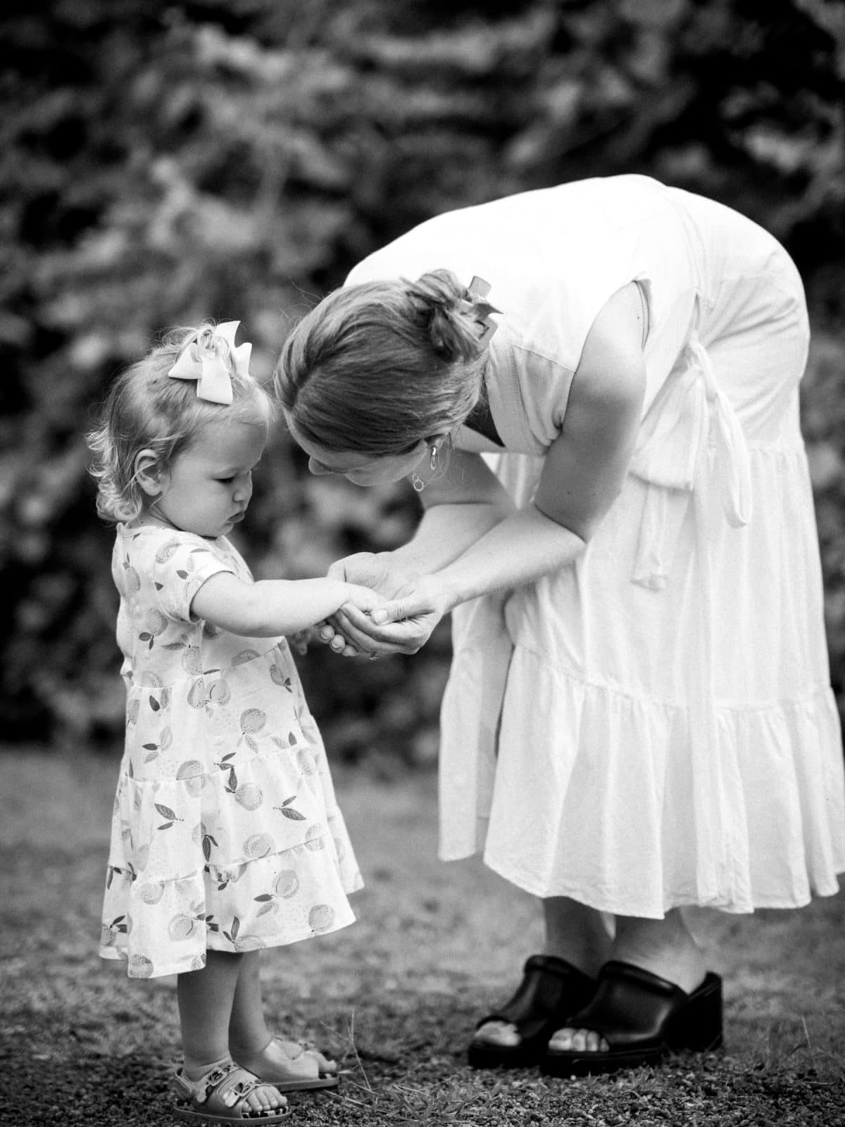 A woman in a white dress bends down to gently talk to a young girl in a patterned dress with bows in their hair. Captured in classic black and white, this beautiful moment of family photography unfolds outdoors, with lush foliage serving as the serene backdrop.