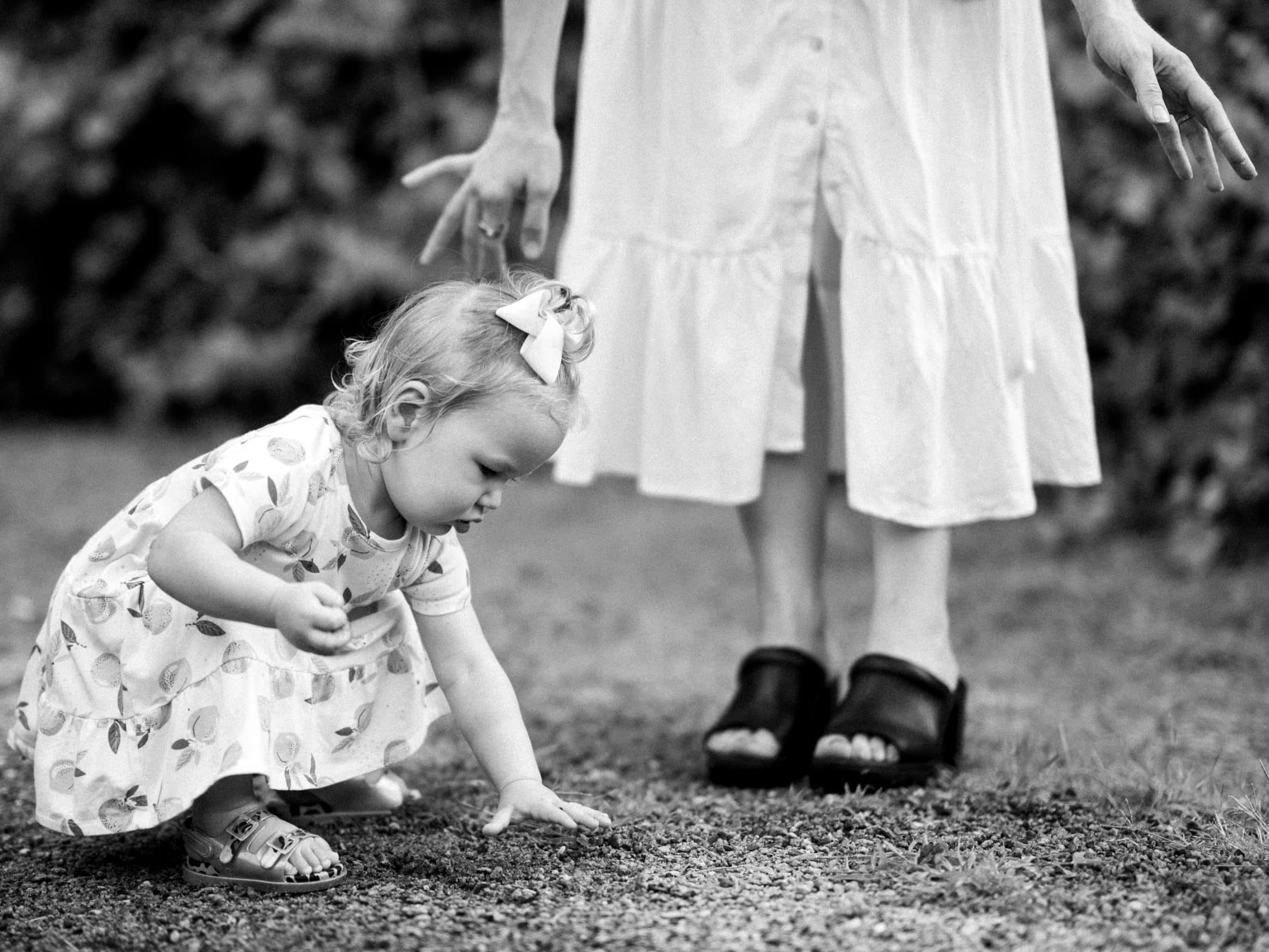 A toddler in a patterned dress and sandals bends down to touch the ground, capturing a candid moment of family photography. An adult in a white dress and black sandals stands nearby, set against a backdrop of blurred greenery in the outdoor scene.