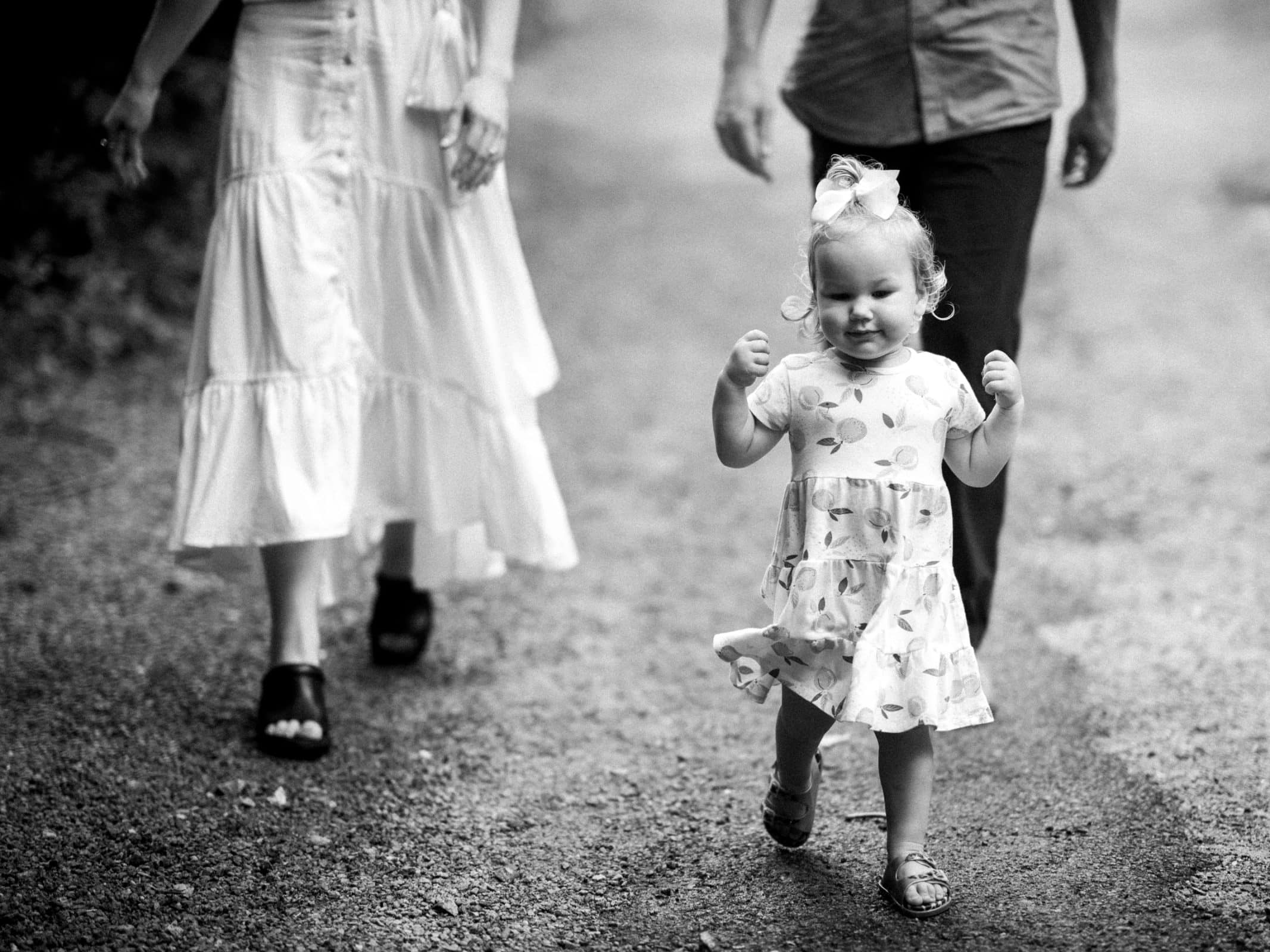 Black and white image of a happy toddler in a dress and bow walking on a path. Two adults, one in a white skirt and another in pants, follow her joyfully. The scene captures the candid essence of family photography.