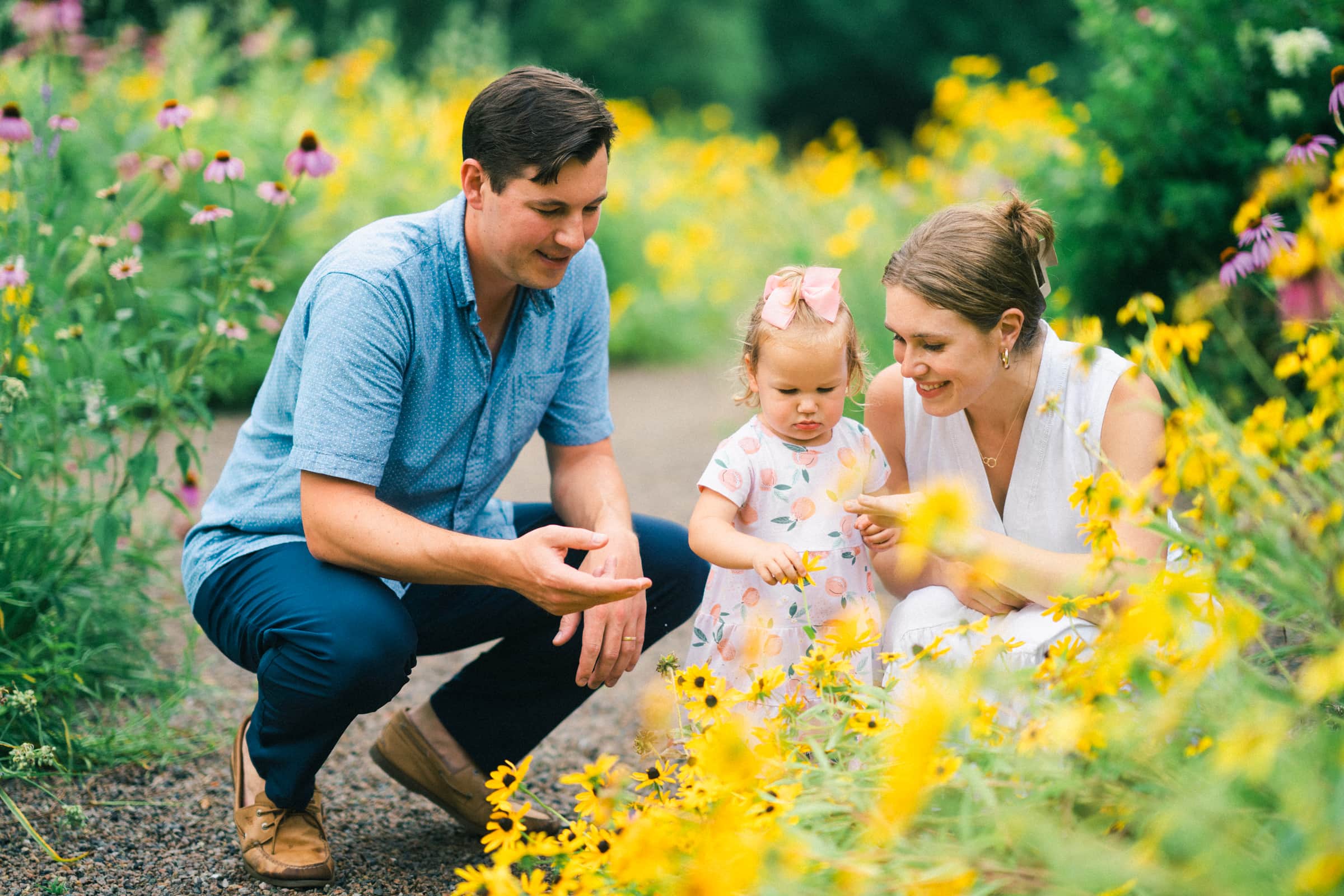 A family of three, captured in a delightful moment of family photography, admire yellow flowers in a garden. The young girl, in a pink bow and floral dress, is flanked by her parents—a man in a blue shirt and a woman in white—surrounded by lush greenery and vibrant blooms.