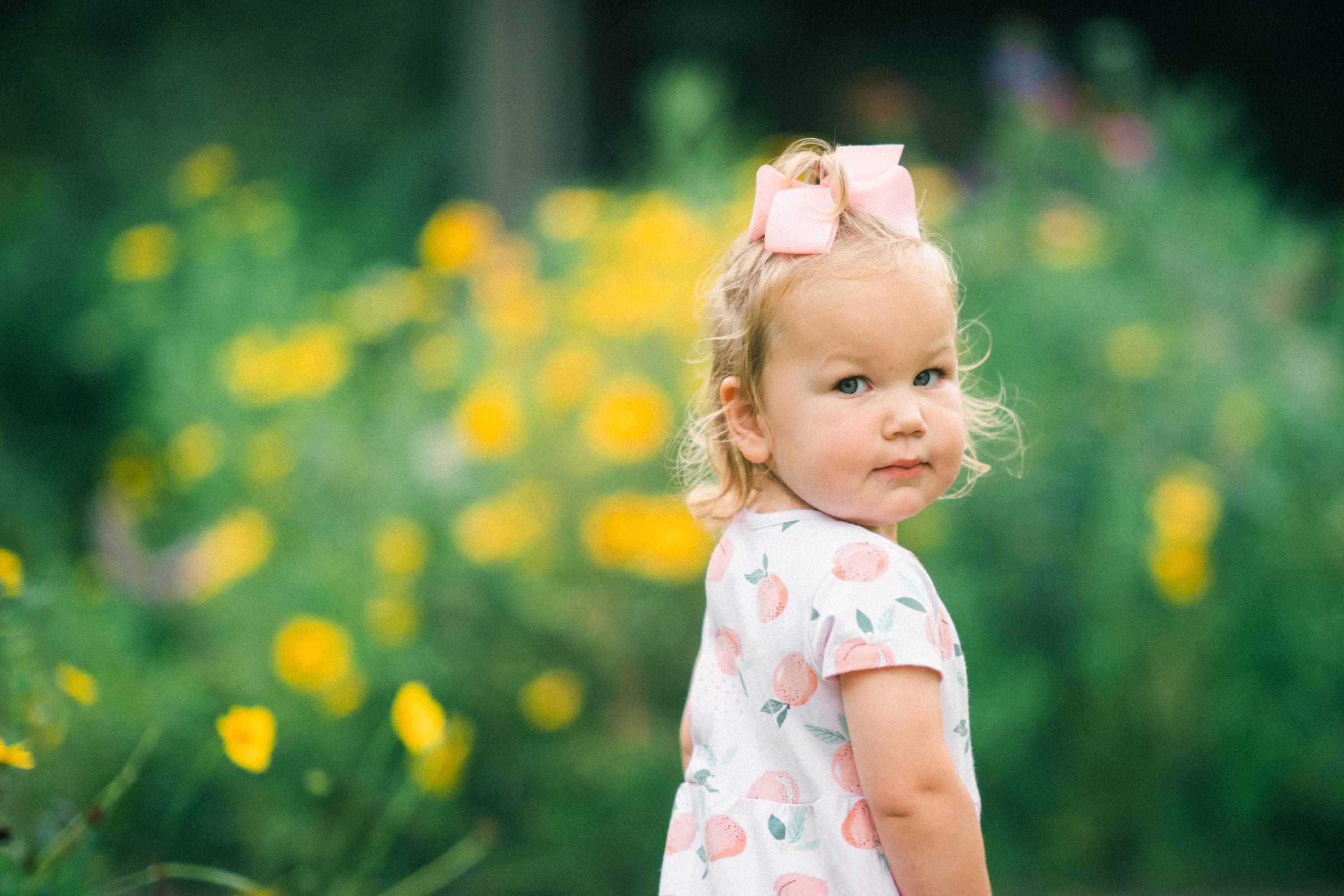 A young girl with a pink bow in her hair stands in a field of yellow flowers, facing to the side, embodying the charm often captured in family photography. She wears a light-colored dress with a peach pattern, set against a lush green background that creates a vibrant and serene setting.