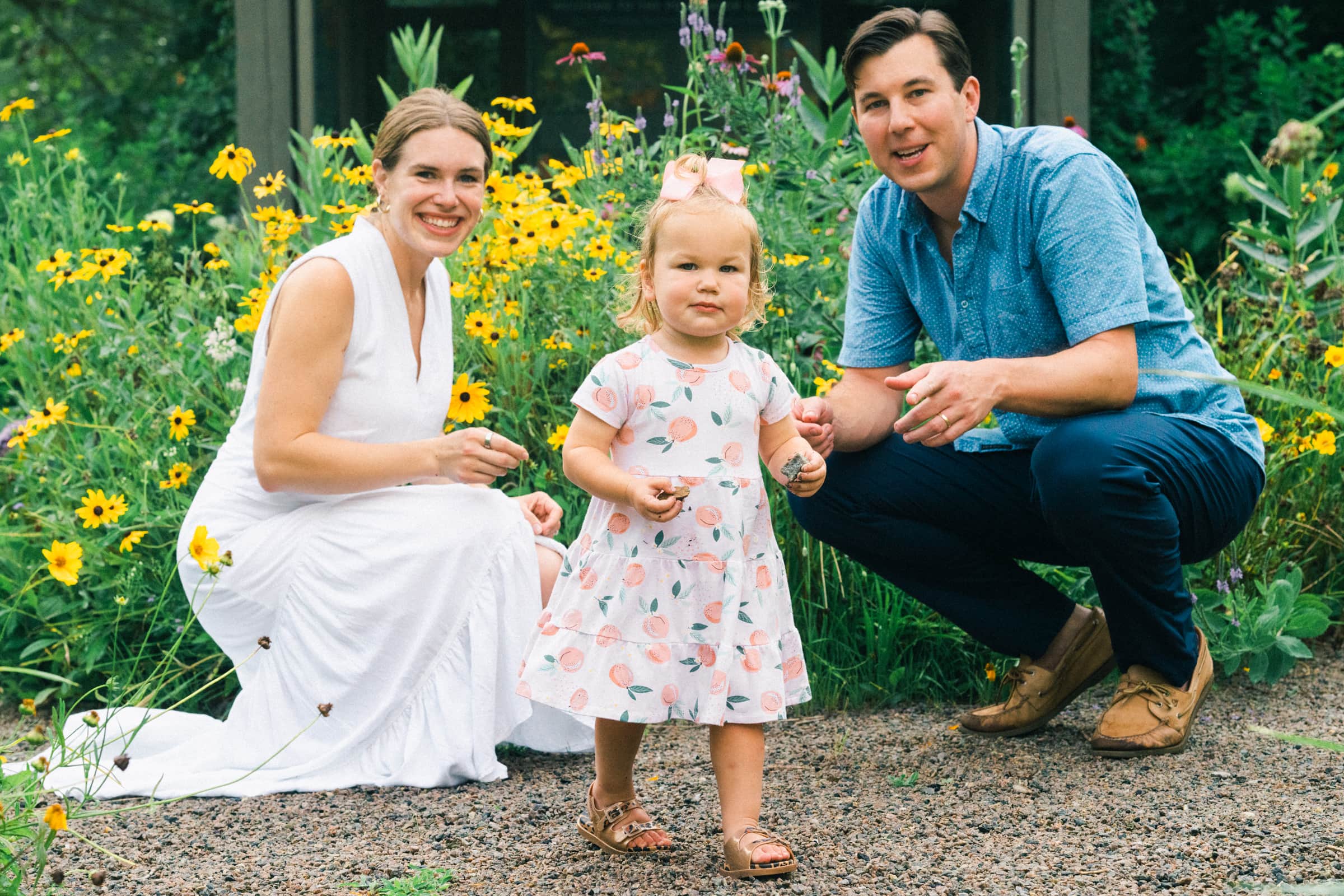 A family of three epitomizes the charm of family photography as they pose in a garden adorned with yellow flowers. The woman in a white dress, the man in a blue shirt, and their toddler, delightfully dressed in floral attire, all smile warmly at the camera while she stands proudly before them.