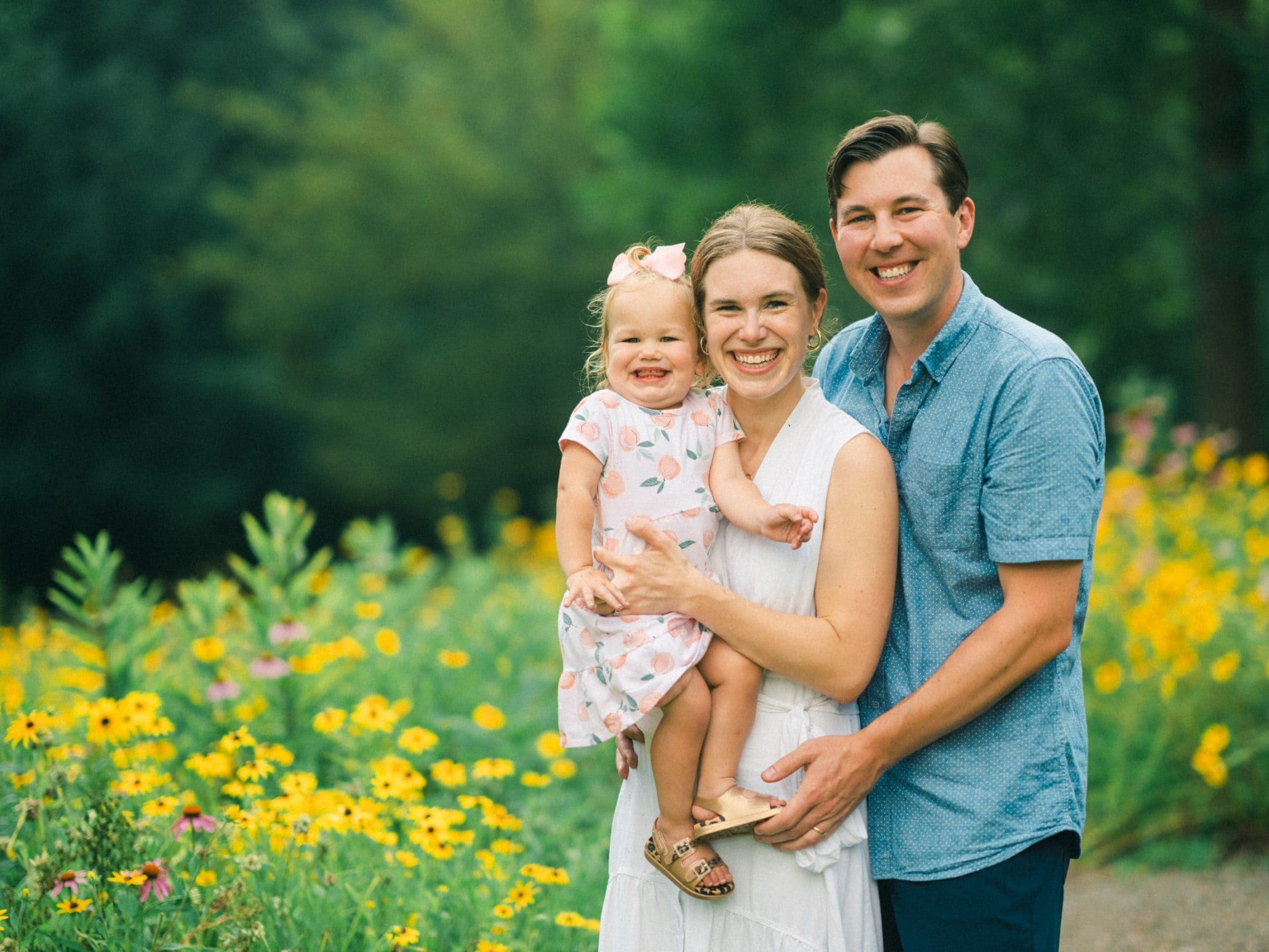 A smiling family of three stands in a field of yellow wildflowers, capturing the essence of family photography. The woman in a white dress, the man in a blue shirt, and their young child in a pink dress all look at the camera, exuding joy and warmth.