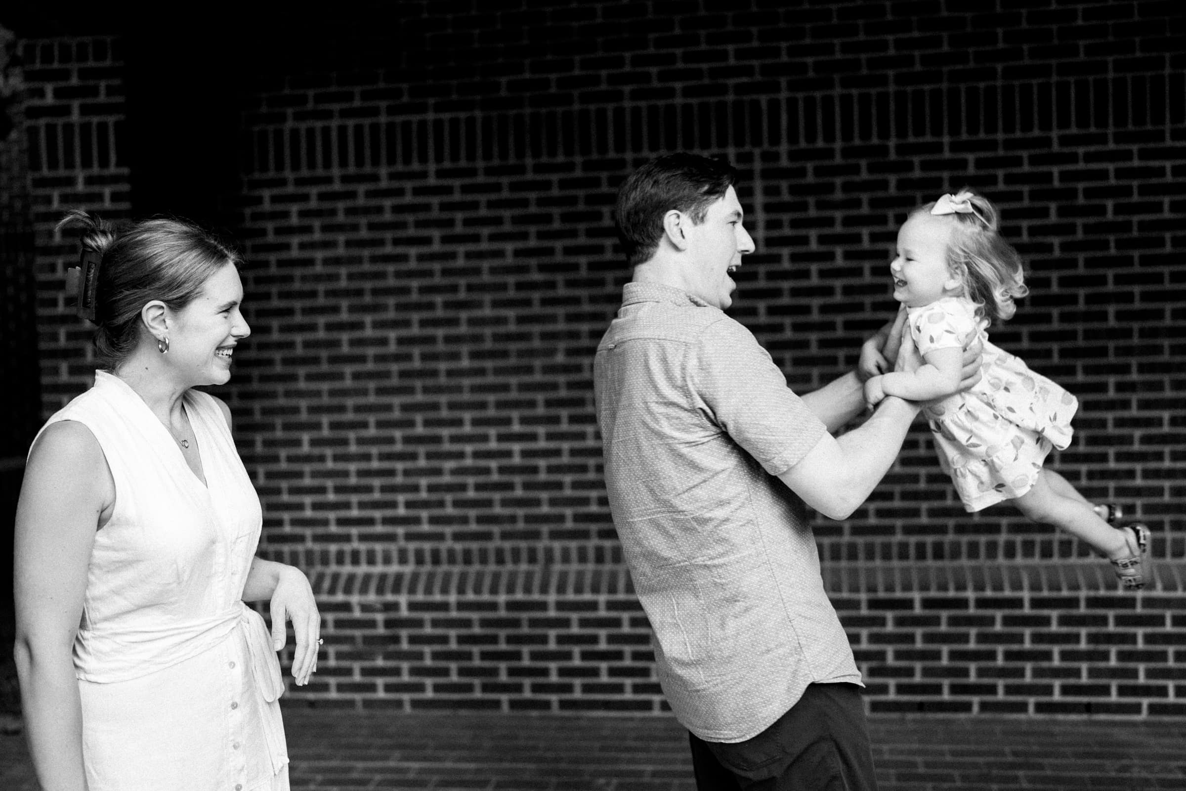 A black and white family photography moment: a man playfully lifts a smiling toddler in a dress, while a woman looks on with delight. They stand joyfully in front of a brick wall.