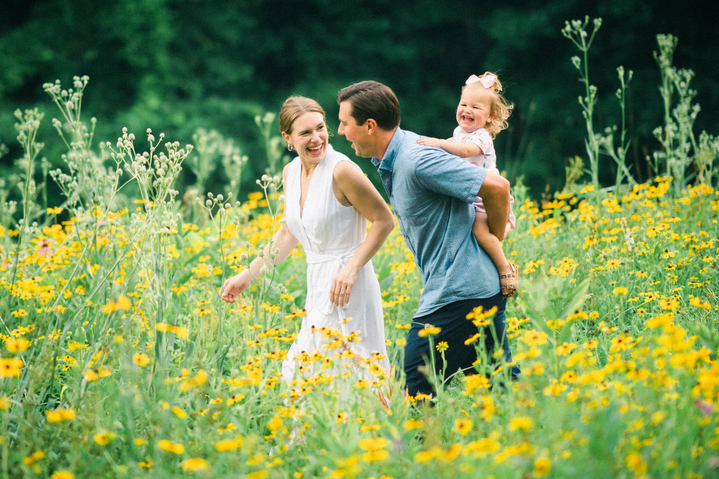 A happy family walks through a vibrant field of yellow wildflowers, perfect for family photography. The woman in a white dress holds hands with a man in a blue shirt, who carries a laughing child on his shoulder. Lush greenery forms the picturesque background.