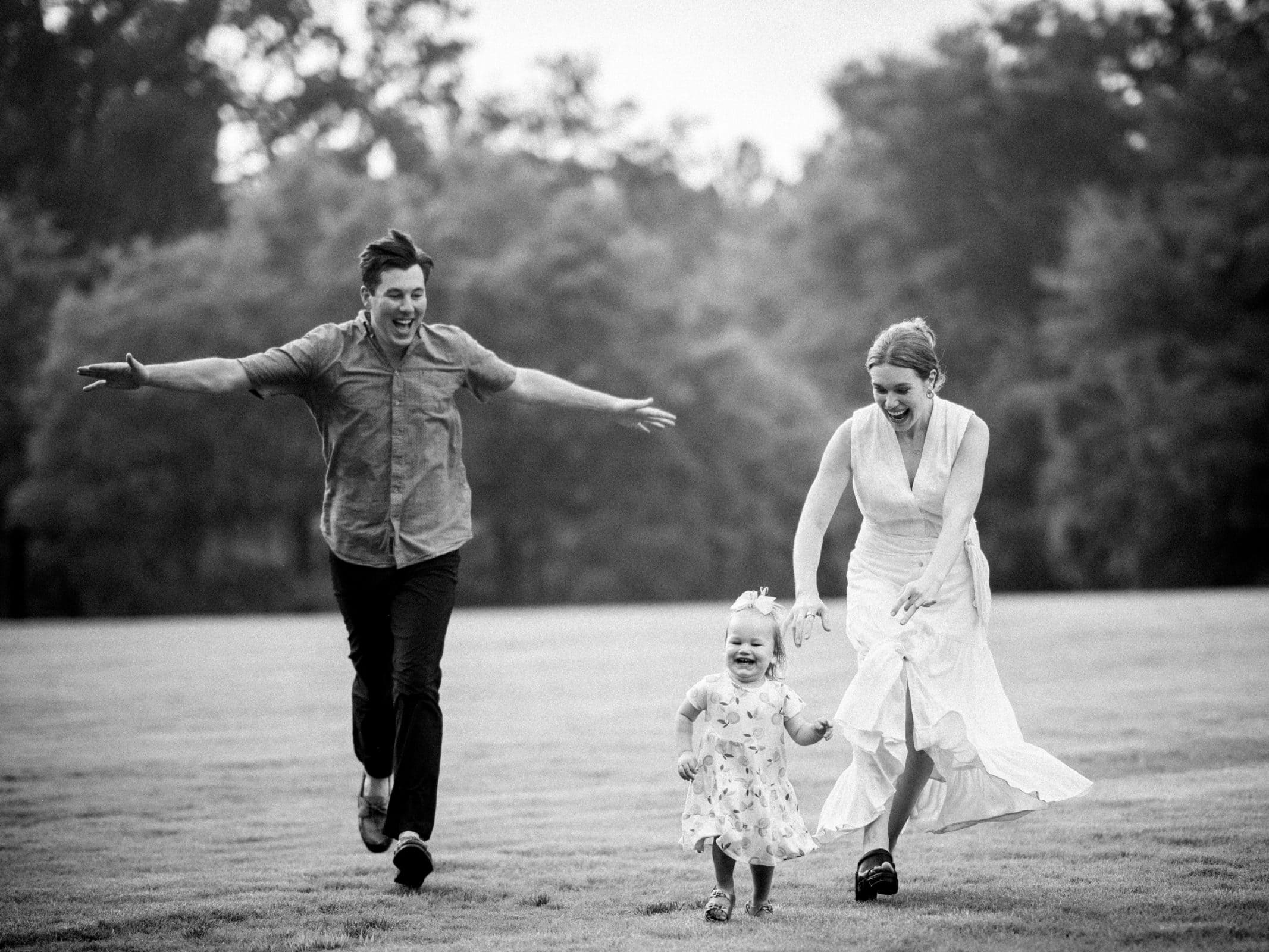 A joyful, black-and-white family photography captures a man and woman playfully running with a toddler across a grassy field. The toddler laughs and reaches forward, while the adults extend their arms wide in happiness. Trees stand silently in the background.