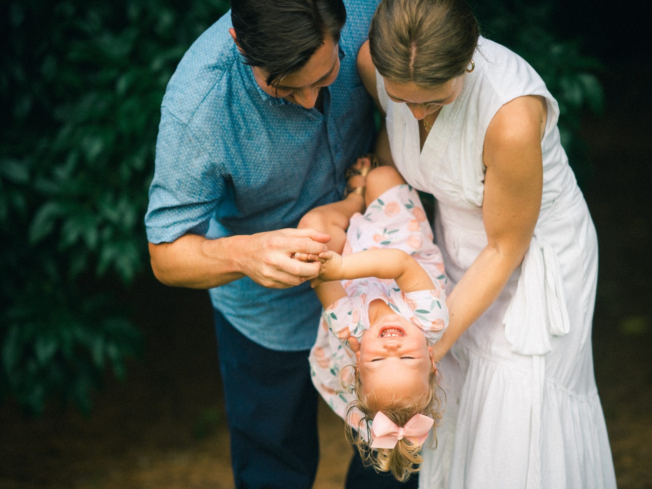 A couple playfully holds a laughing toddler upside down, capturing the essence of family photography. The child, in a floral dress with a pink bow in her hair, giggles as the adults share smiles amidst a backdrop of lush greenery.