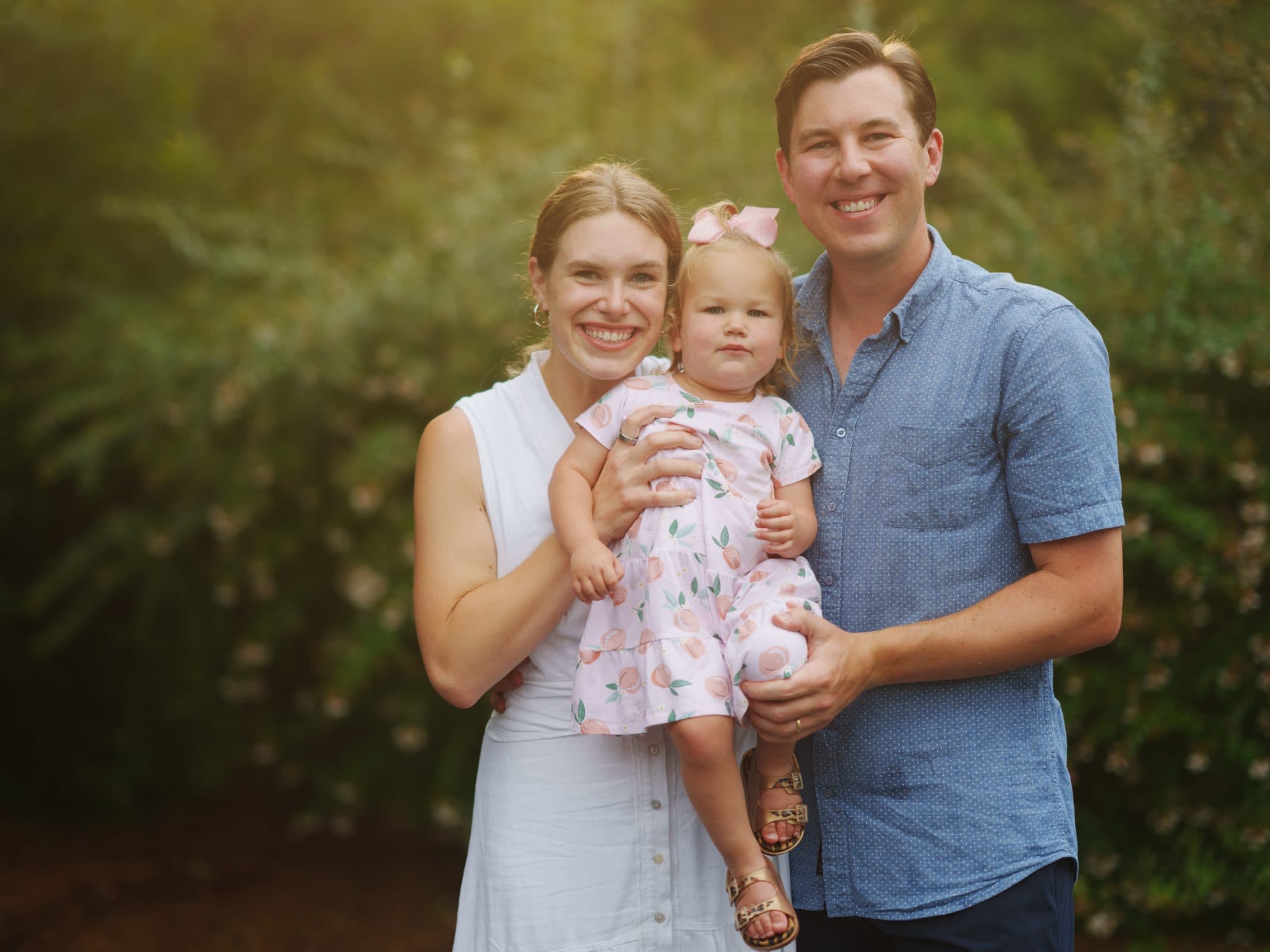A smiling couple stands with a toddler, capturing the essence of family photography. The woman wears a white dress, and the man sports a blue shirt. The child, dressed in a patterned dress with a bow, is held lovingly between them. Greenery fills the background.