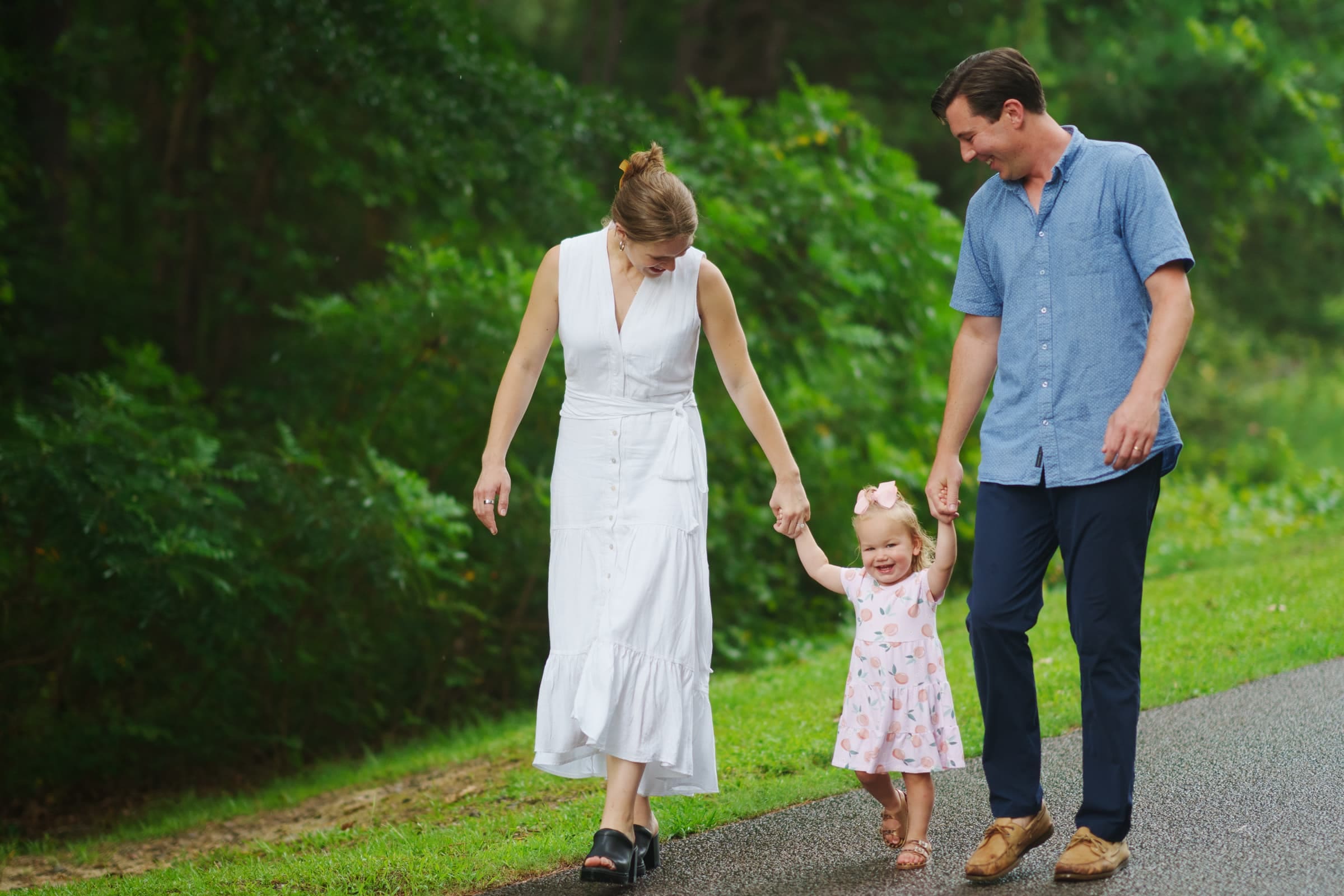 A smiling couple walks hand in hand with their young daughter on a lush green path, capturing a perfect family photography moment. The woman in a white dress and the man in a blue shirt hold the girl's hands as she happily swings in the air.