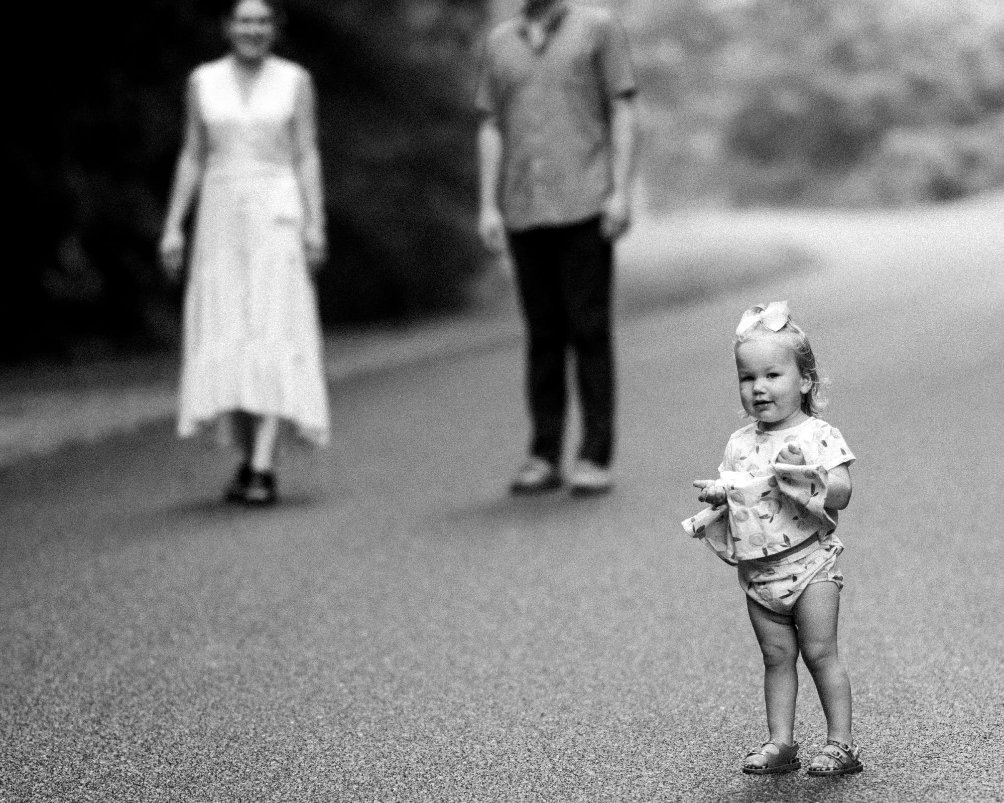 A black and white family photography image captures a toddler standing on a road, dressed in a bow and patterned outfit. In the blurred background, two adults approach: one in a dress and the other in pants and a shirt.