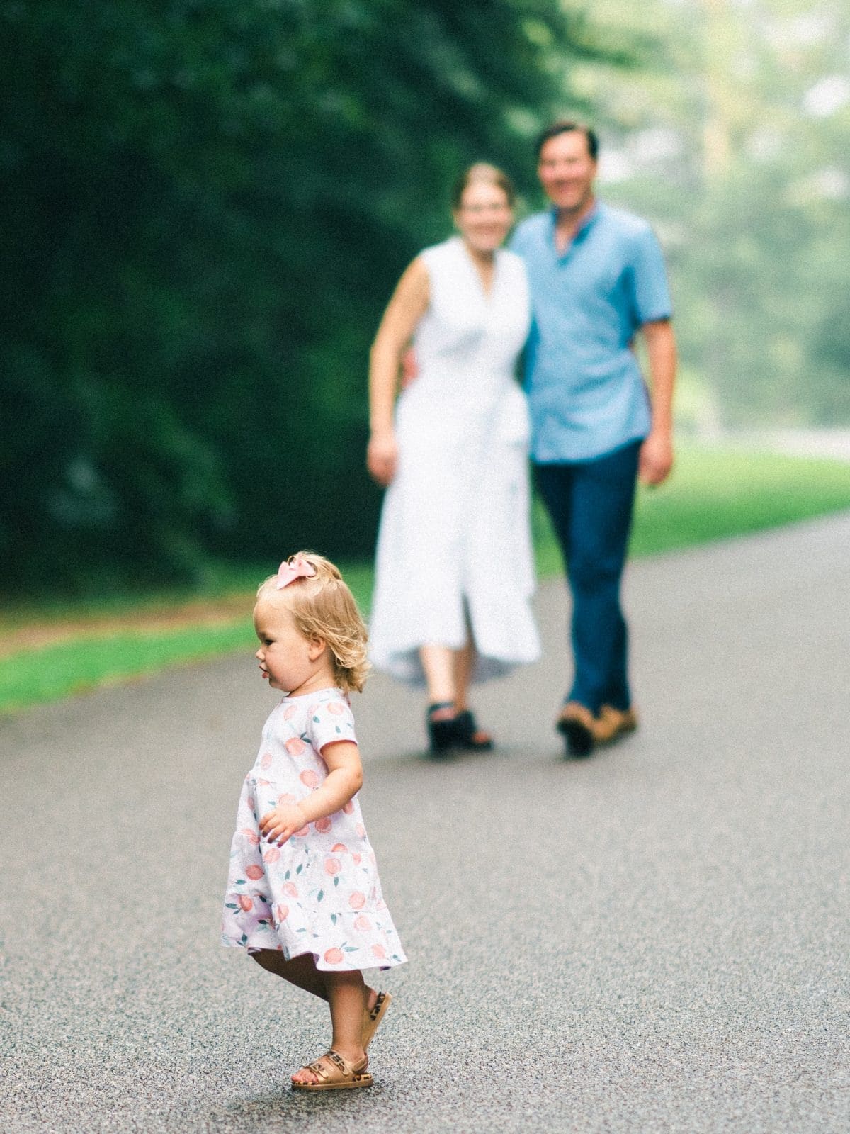 A small child in a patterned dress walks on a paved path, capturing the essence of family photography. In the background, slightly out of focus, two adults stroll together amidst lush greenery, evoking a serene park or garden setting.
