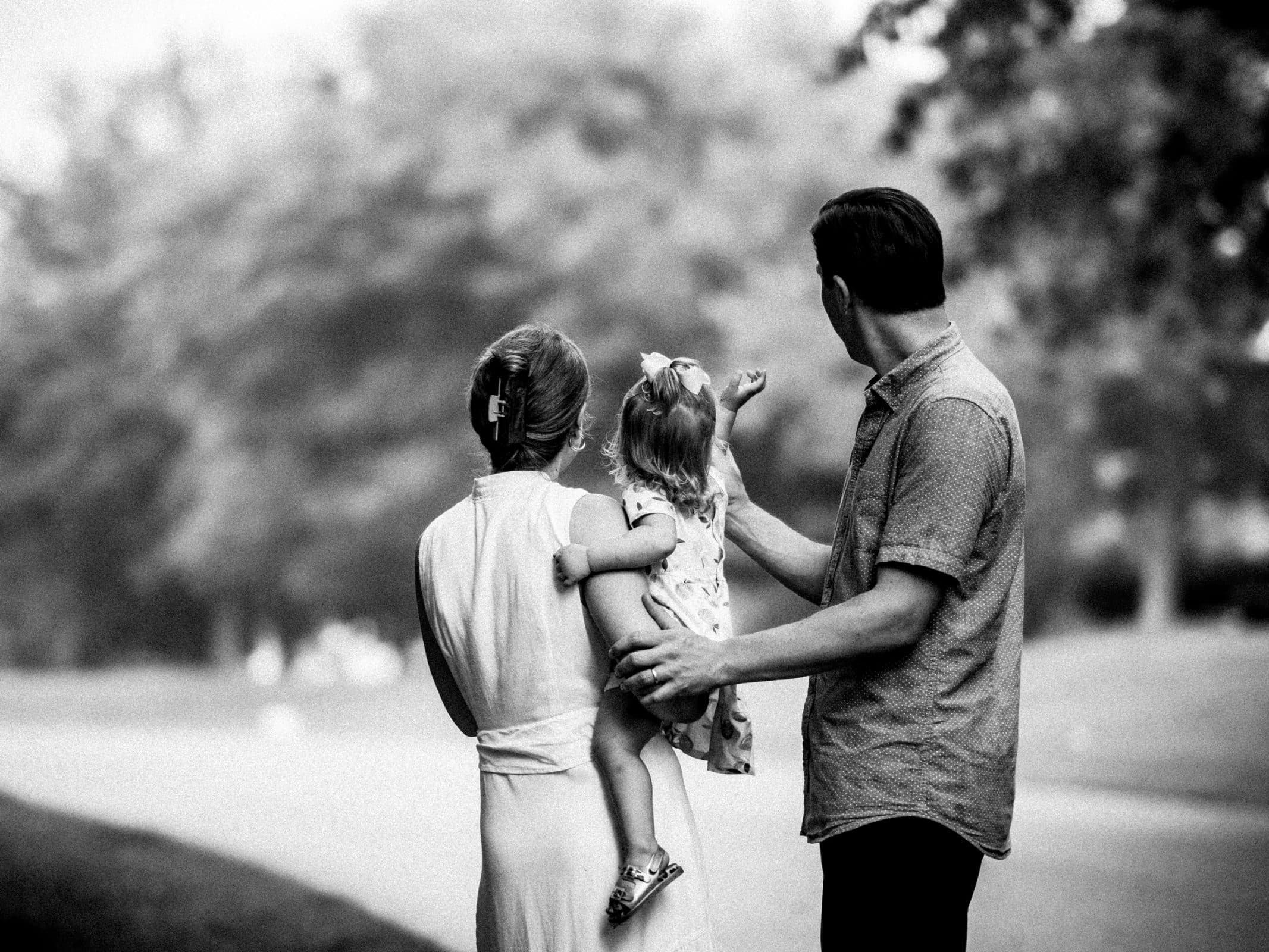 Black and white family photography captures a couple with their child. The woman, in a dress, holds the little girl adorned with a bow in her hair. The man points to something in the distance as they stand outdoors, enveloped by trees.