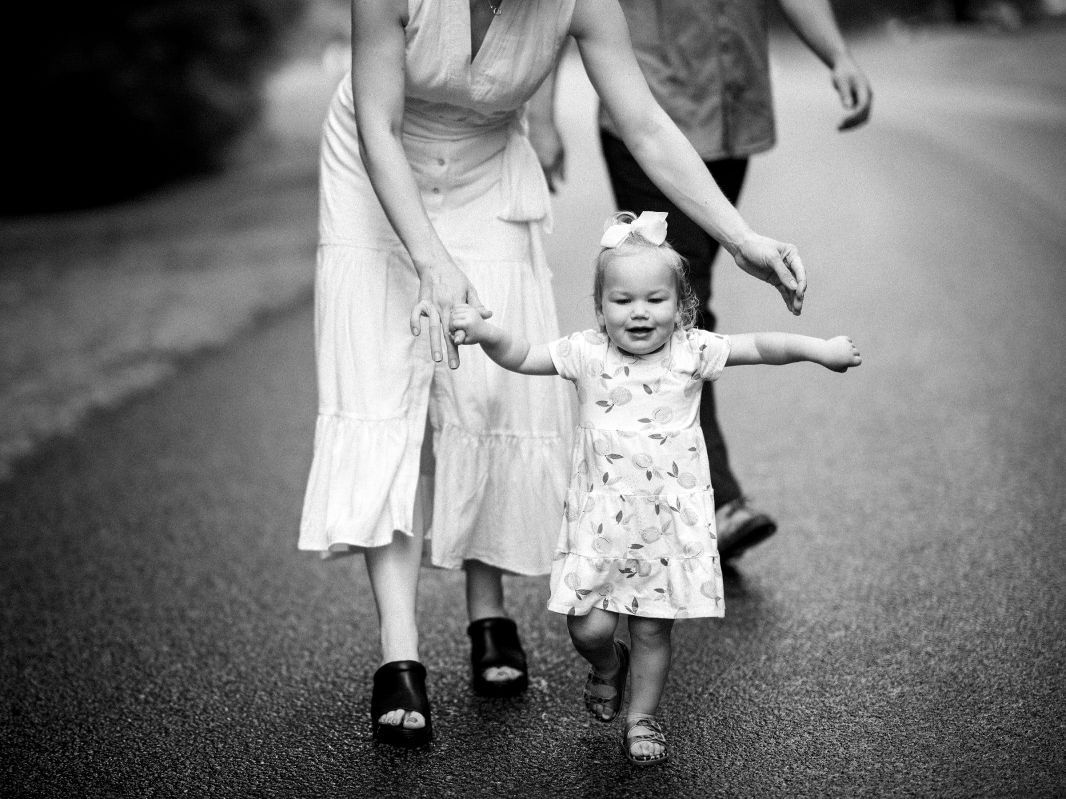 A black and white family photography capture of a young child joyfully walking on a road, supported by guiding hands. The child wears a dress with a bow in their hair, smiling as two adults linger partially visible in the background.