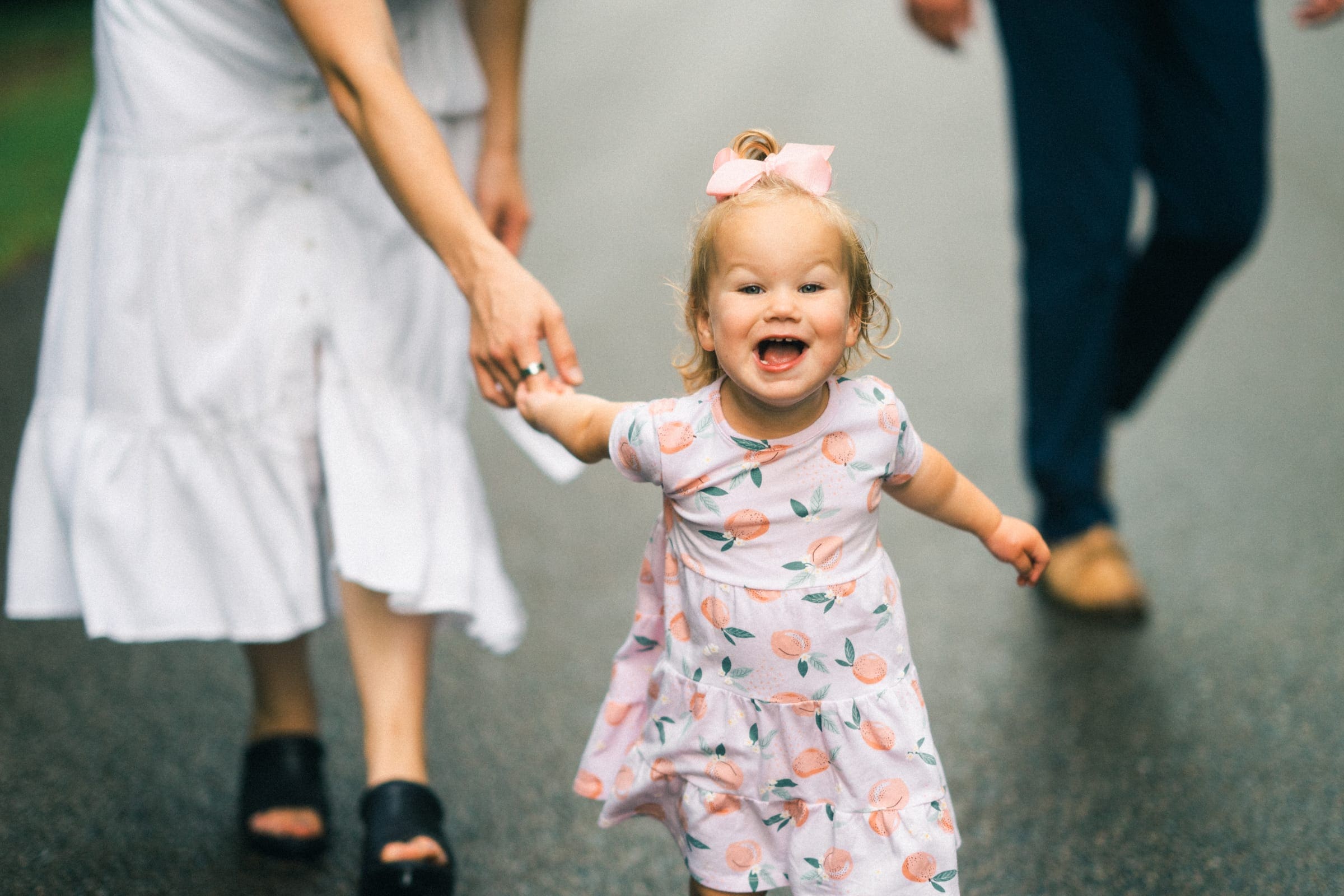A cheerful toddler in a peach-patterned dress and pink bow runs toward the camera, embodying the joy of family photography. She holds the hand of an adult in a white dress, while another adult in blue pants and brown shoes follows closely on the paved path.